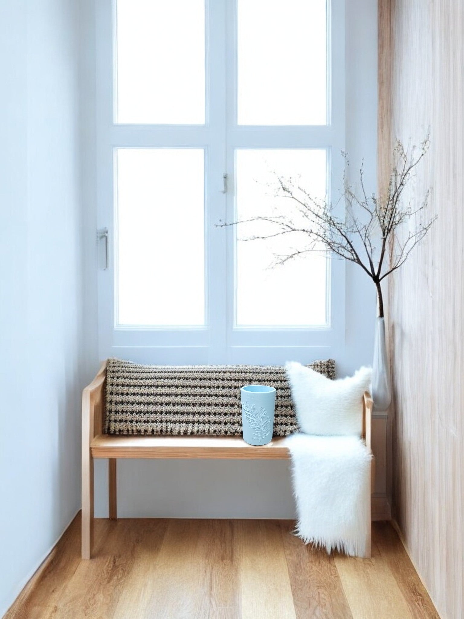 A minimalist seating nook features a wooden bench, textured cushion, white faux fur pillow, blue cup, and the Glacial Blue Winter Fern Vase – Woodland Accent by Mirabilia Boutique with bare branches, set against large windows and light wood floors.