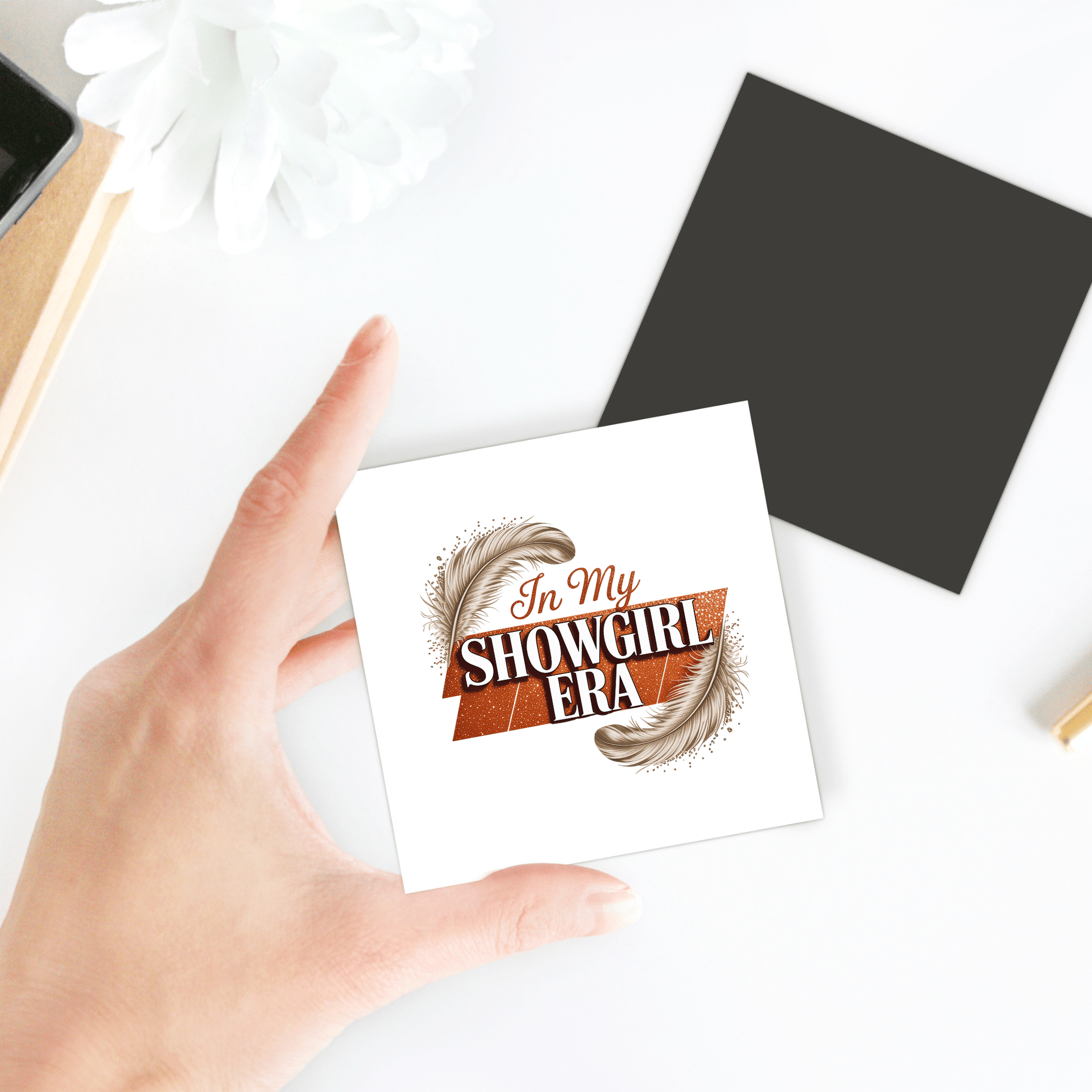 A hand holds the "In My Showgirl Era Magnet" by Mirabilia Boutique—glam glitter vibes for fun fridge decor. The magnet, accented with two feathers, sits on a white surface near a flower and office supplies.