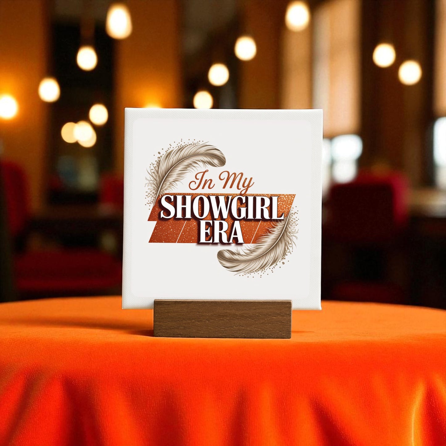 The "In My Showgirl Era Mini Sign" by Mirabilia Boutique adds retro glam to an orange table, featuring bold lettering and feathers, in a warmly lit room with blurred lights and red chairs in the background.