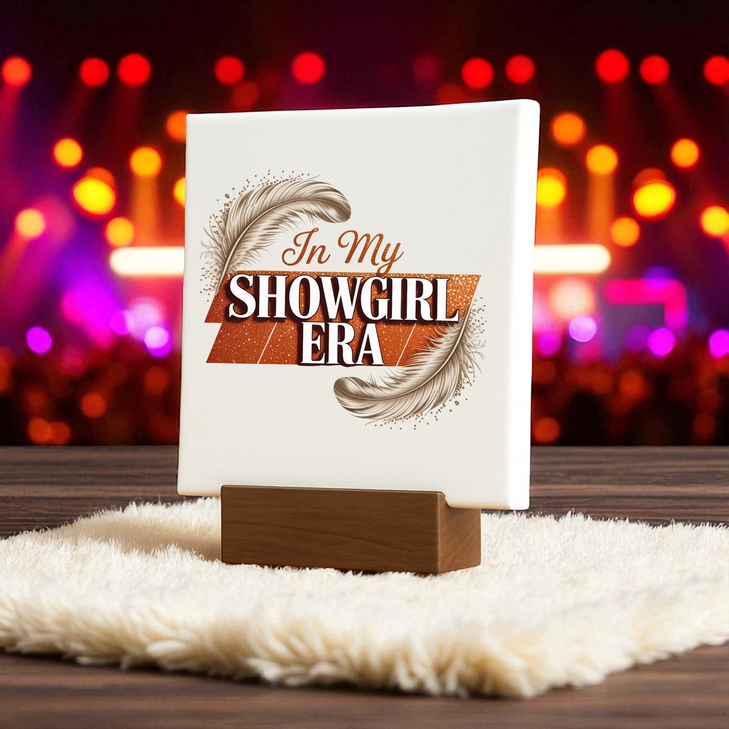The "In My Showgirl Era Mini Sign" by Mirabilia Boutique stands on a wooden base with feather accents, atop a fluffy mat. Blurred colorful stage lights in the background create retro glam decor vibes.