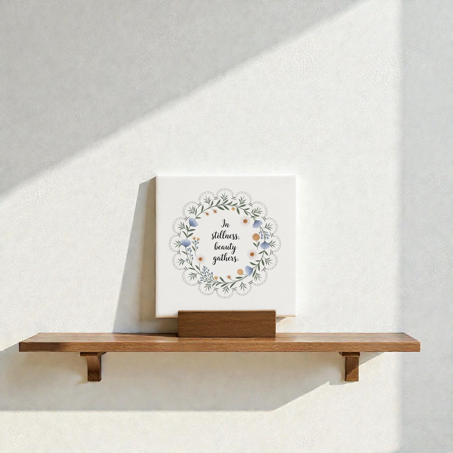 The Mirabilia Boutique "In Stillness Beauty Gathers" Mini Canvas Sign, featuring floral designs, rests on a wooden shelf against a sunlit wall—an ideal meditation decor accent.