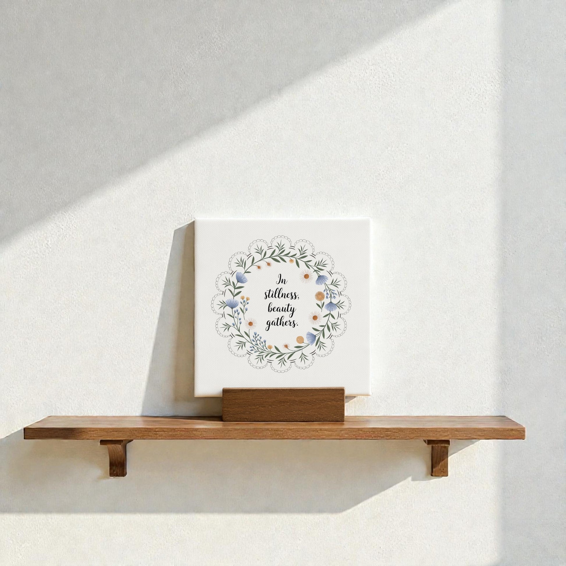 The Mirabilia Boutique "In Stillness Beauty Gathers" Mini Canvas Sign, featuring floral designs, rests on a wooden shelf against a sunlit wall—an ideal meditation decor accent.