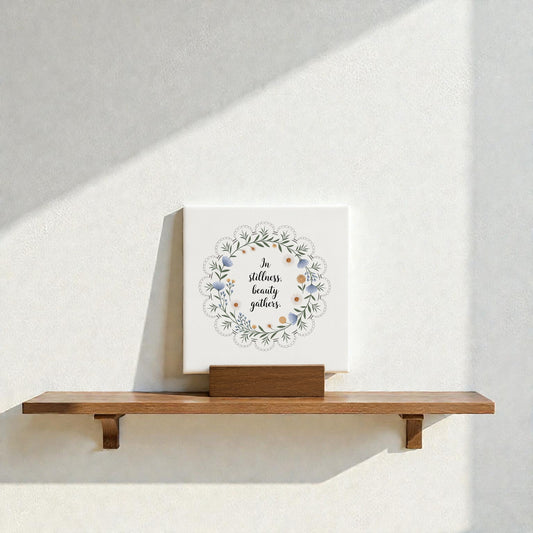 The Mirabilia Boutique "In Stillness Beauty Gathers" Mini Canvas Sign, featuring floral designs, rests on a wooden shelf against a sunlit wall—an ideal meditation decor accent.