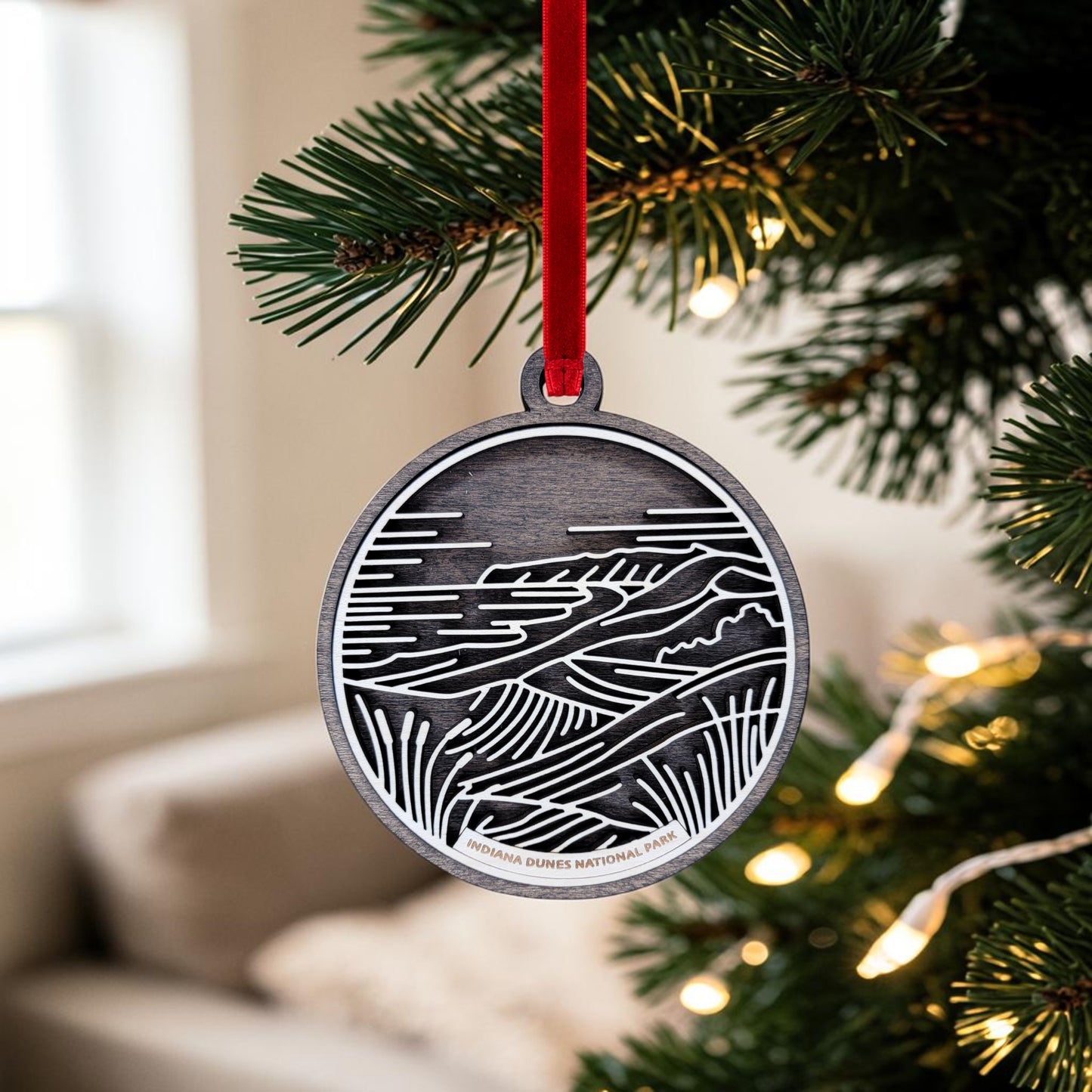 The Indiana Dunes National Park Line Art Christmas Ornament by Mirabilia Boutique, a decorative oval wooden piece adorned with a landscape design inspired by Indiana Dunes National Park, hangs from a red ribbon on a Christmas tree branch amidst soft lights and a softly blurred indoor background.