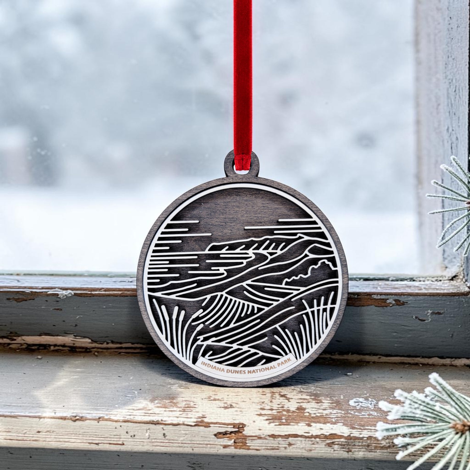 The Indiana Dunes National Park Line Art Christmas Ornament by Mirabilia Boutique—a minimalist oval wooden keepsake—features a landscape design inspired by the park, hanging from a red ribbon on a window latch; with frosted glass and a pine branch in the background, it creates a wintry Lake Michigan scene.