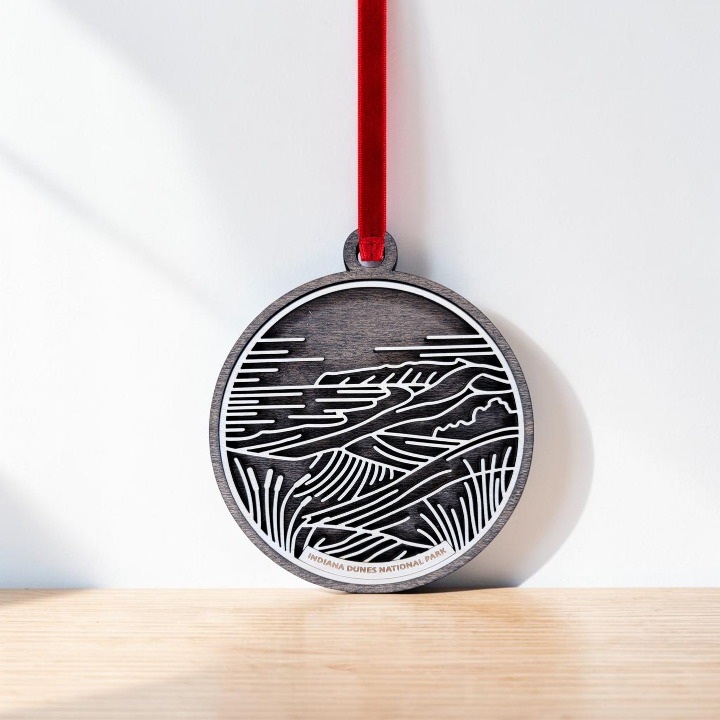 The Indiana Dunes National Park Line Art Christmas Ornament by Mirabilia Boutique features a round wooden design with light line art depicting a stylized landscape. Hanging from a red ribbon, this ornament makes an ideal Lake Michigan keepsake, with “Indiana Dunes National Park” inscribed at the bottom.