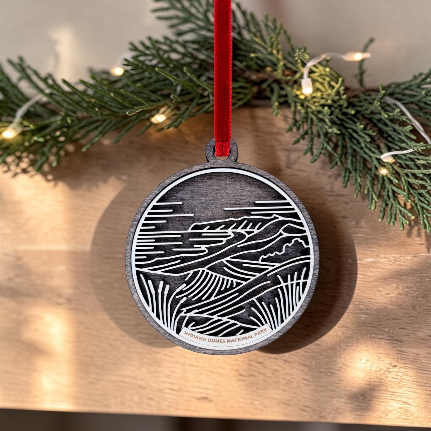 The Indiana Dunes National Park Line Art Christmas Ornament by Mirabilia Boutique is an oval wooden ornament featuring a landscape design and the text "Indiana Dunes National Park" at the bottom. It hangs from a red ribbon in front of pine branches and string lights.