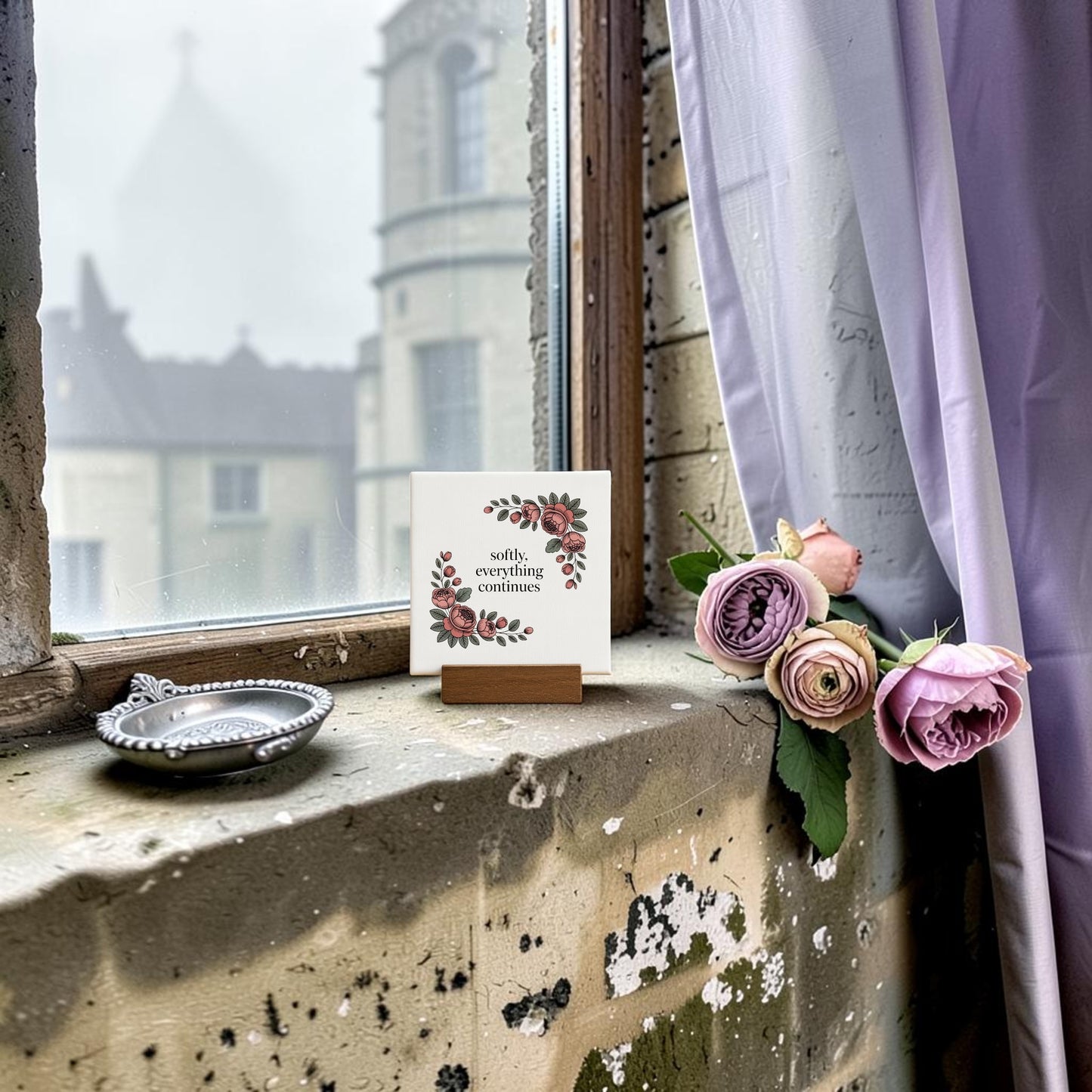 A weathered windowsill with pink roses, a silver dish, and the Mirabilia Boutique Softly, Everything Continues Mini Sign—an elegant decor piece and thoughtful gift. Outside, a stone building is visible and a lavender curtain drapes to the right.