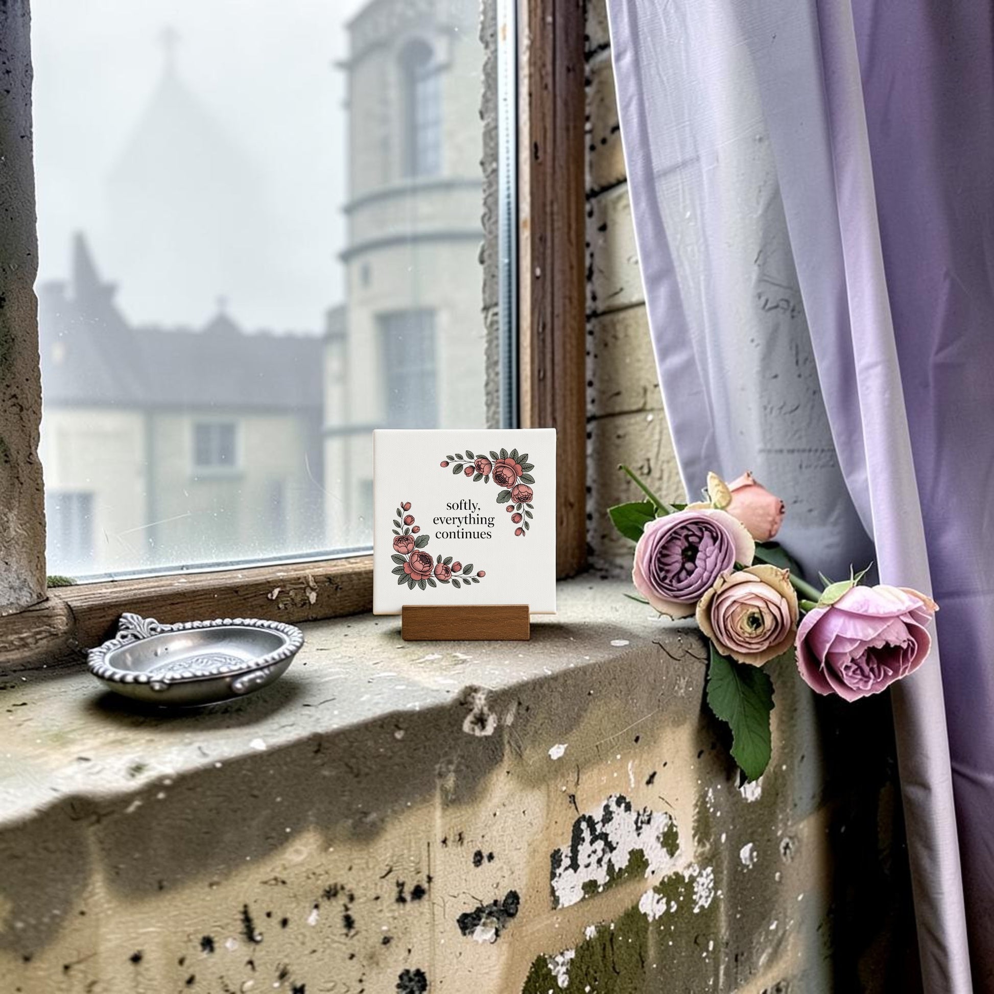 A weathered windowsill with pink roses, a silver dish, and the Mirabilia Boutique Softly, Everything Continues Mini Sign—an elegant decor piece and thoughtful gift. Outside, a stone building is visible and a lavender curtain drapes to the right.