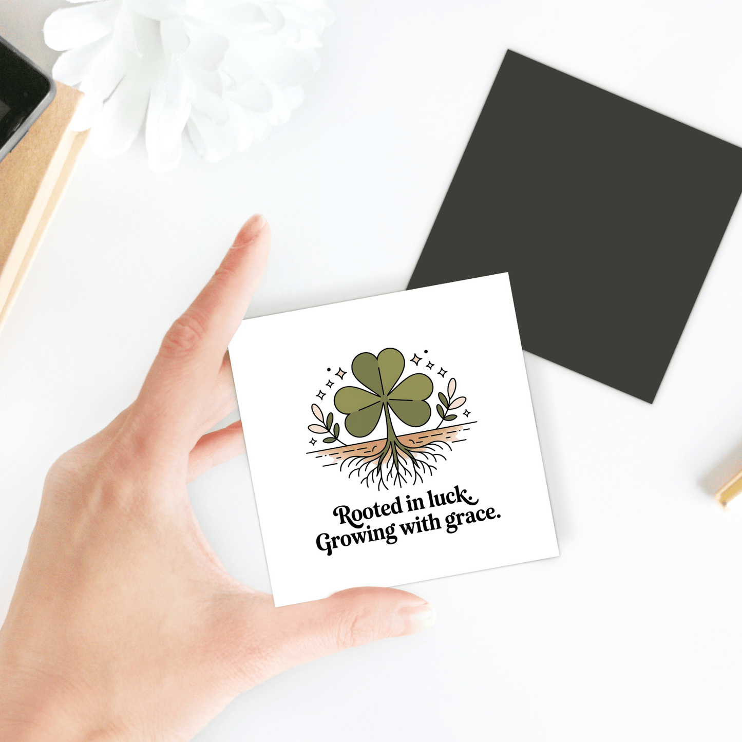 A hand holds the Mirabilia Boutique "Rooted in Luck Shamrock Magnet," featuring a clover with visible roots and the phrase "Rooted in luck. Growing with grace." A white flower and black square sit on a white surface in the background.