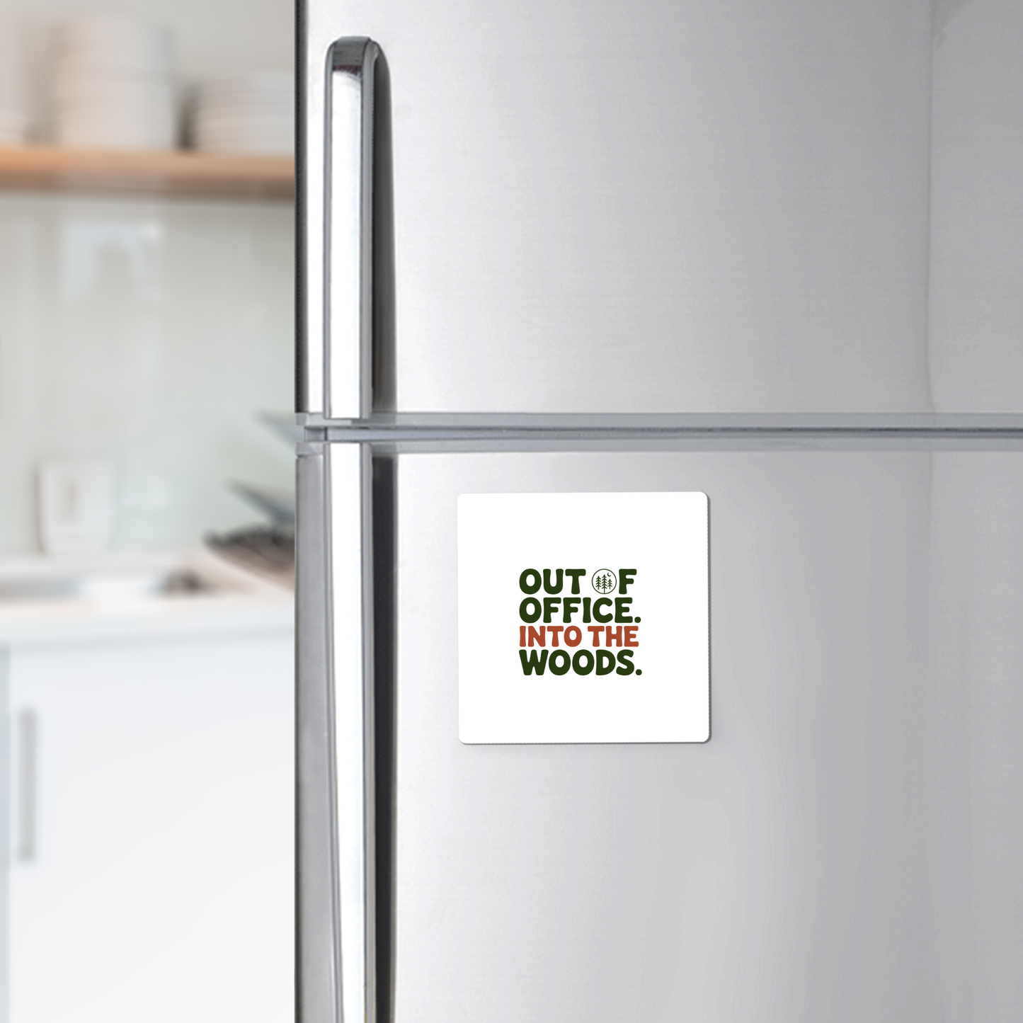 The Out of Office Into the Woods Magnet by Mirabilia Boutique—a square white magnet with green text—is shown on a stainless steel fridge, perfect as cabin decor or a gift for nature lovers. White dishes appear blurred in the kitchen background.