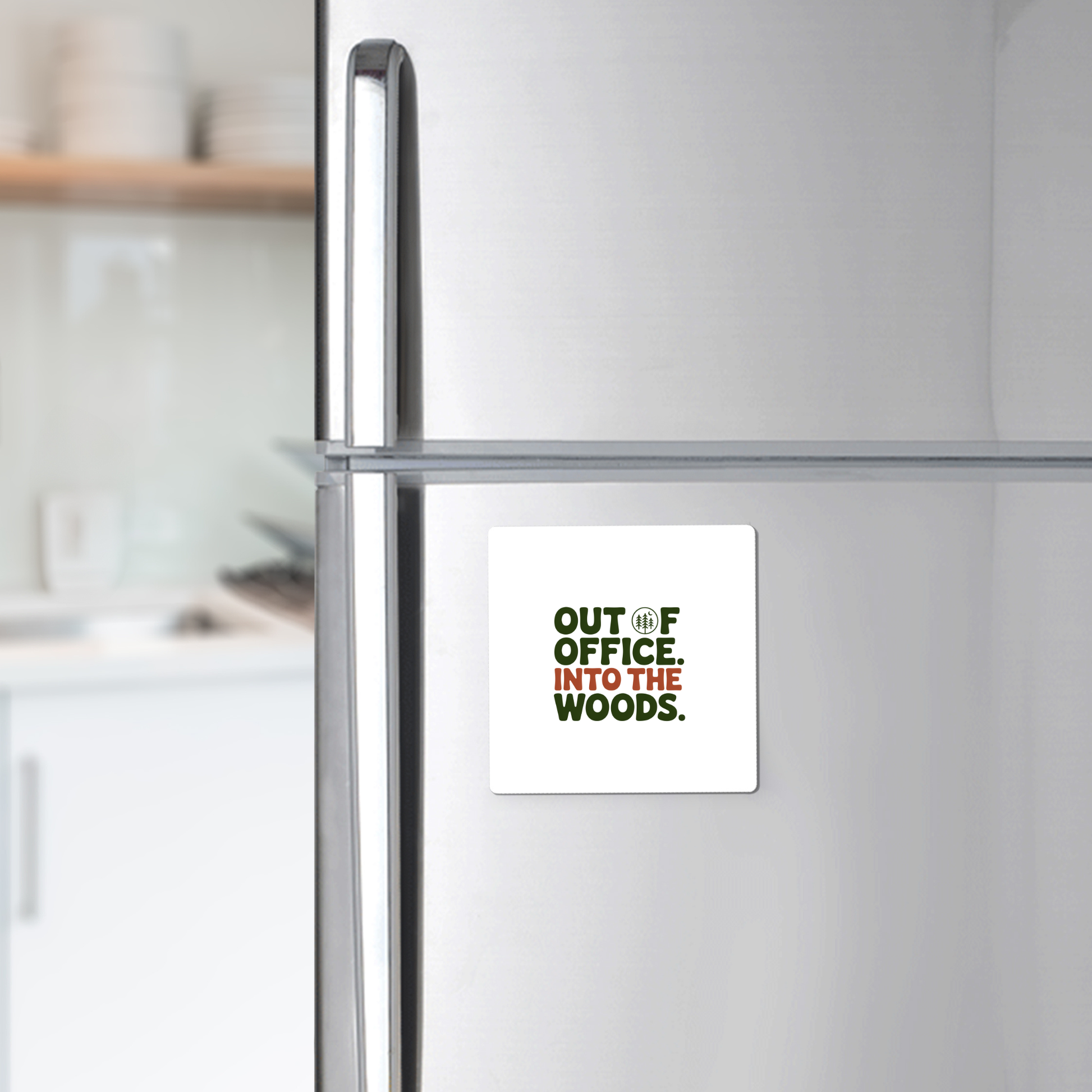 The Out of Office Into the Woods Magnet by Mirabilia Boutique—a square white magnet with green text—is shown on a stainless steel fridge, perfect as cabin decor or a gift for nature lovers. White dishes appear blurred in the kitchen background.