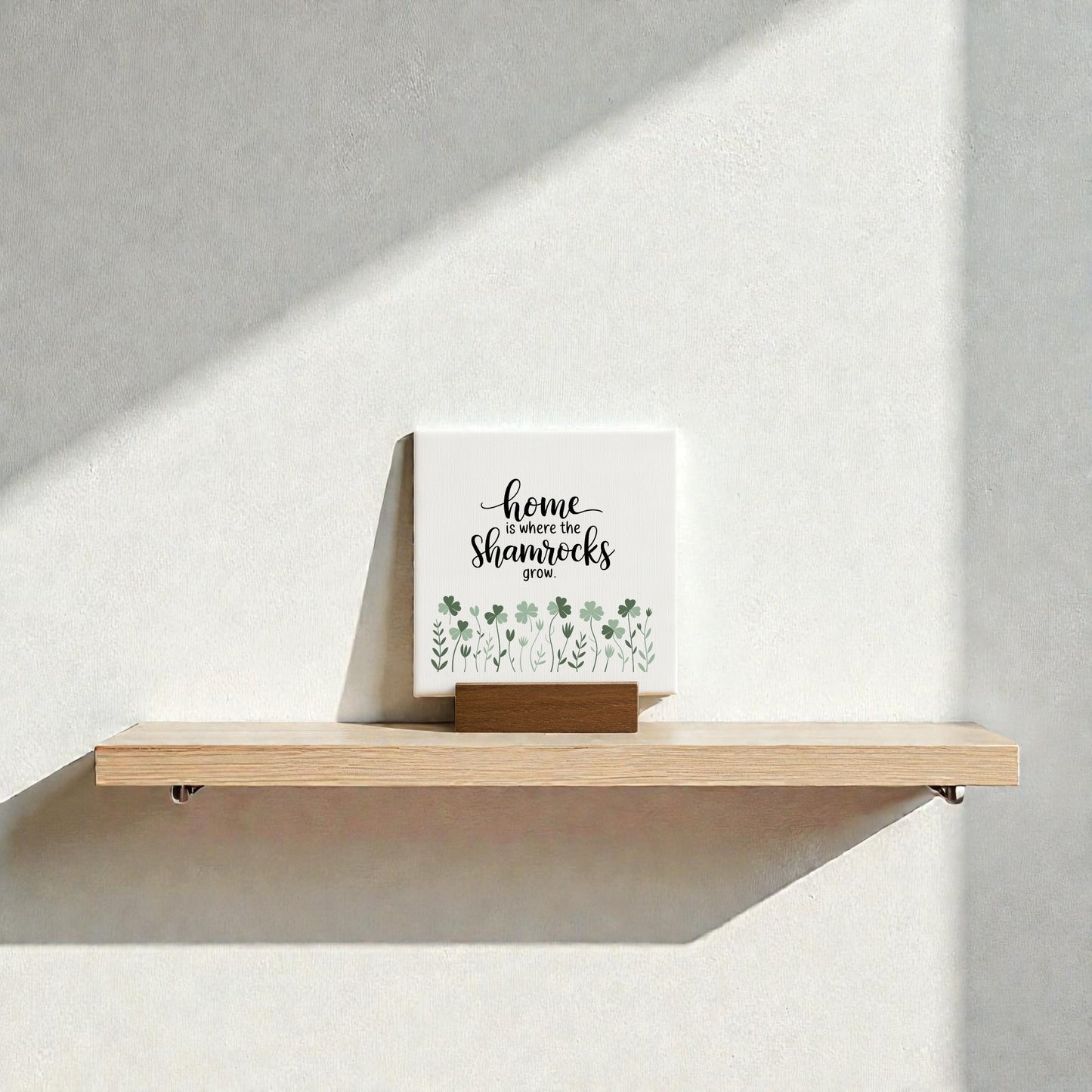 A Mirabilia Boutique "Home Is Where the Shamrocks Grow" mini sign with green shamrock art sits on a wooden shelf against a white wall, sunlight casting gentle shadows for charming Irish home decor.