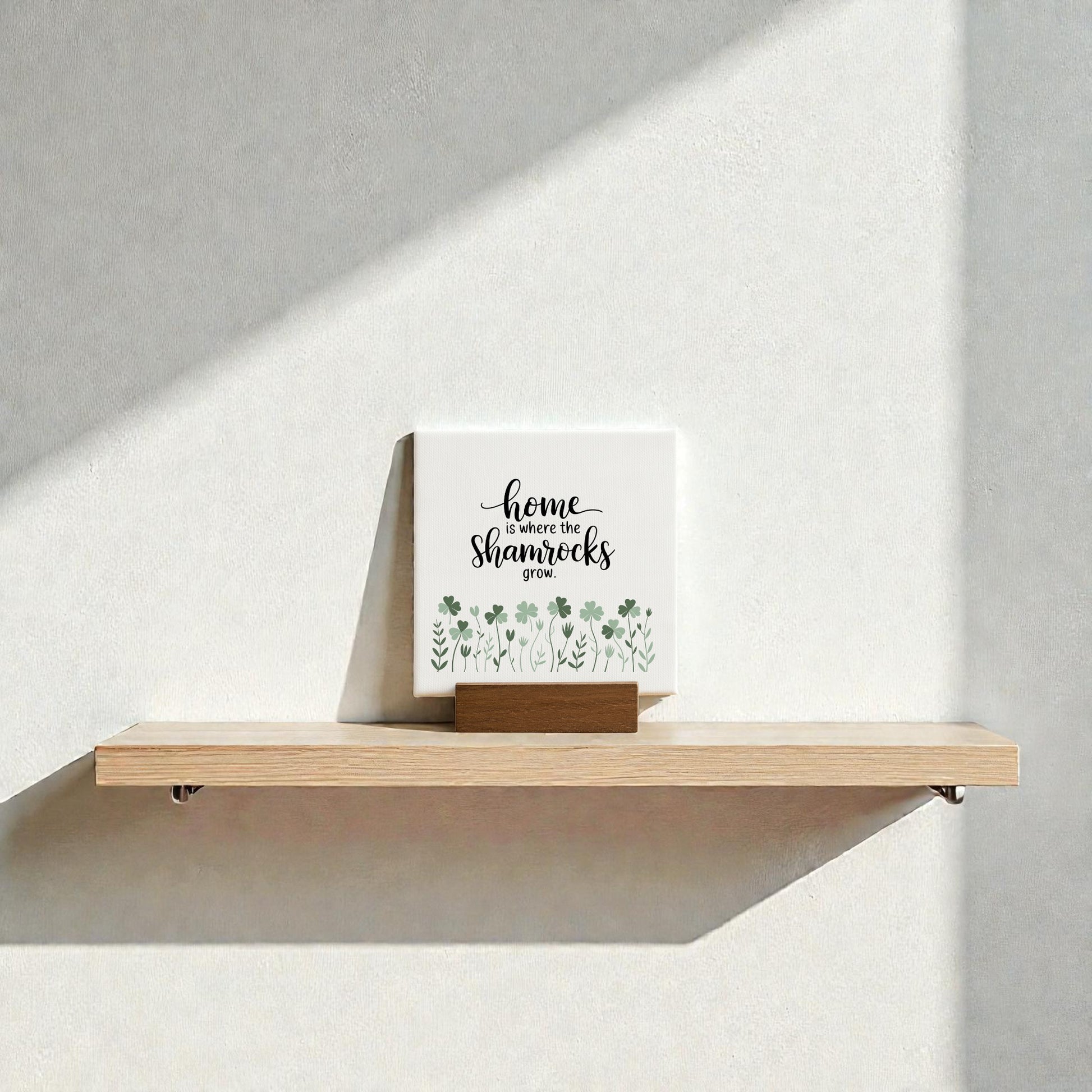 A Mirabilia Boutique "Home Is Where the Shamrocks Grow" mini sign with green shamrock art sits on a wooden shelf against a white wall, sunlight casting gentle shadows for charming Irish home decor.
