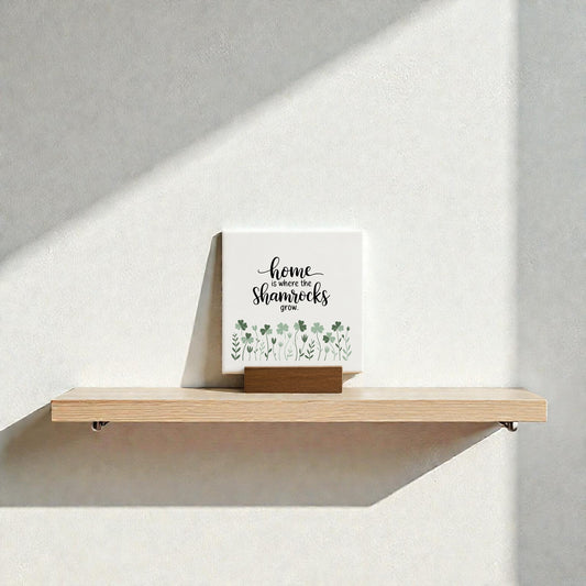 A Mirabilia Boutique "Home Is Where the Shamrocks Grow" mini sign with green shamrock art sits on a wooden shelf against a white wall, sunlight casting gentle shadows for charming Irish home decor.