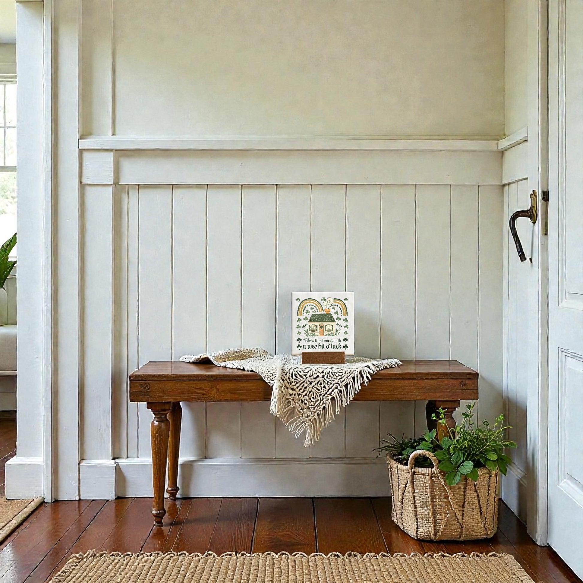 A wooden bench with a beige lace cloth, Mirabilia Boutique's "A Wee Bit of Luck Canvas Sign – Irish Cottage Charm," and a woven basket of greenery sits against a white paneled wall in a bright entryway with hardwood floors.