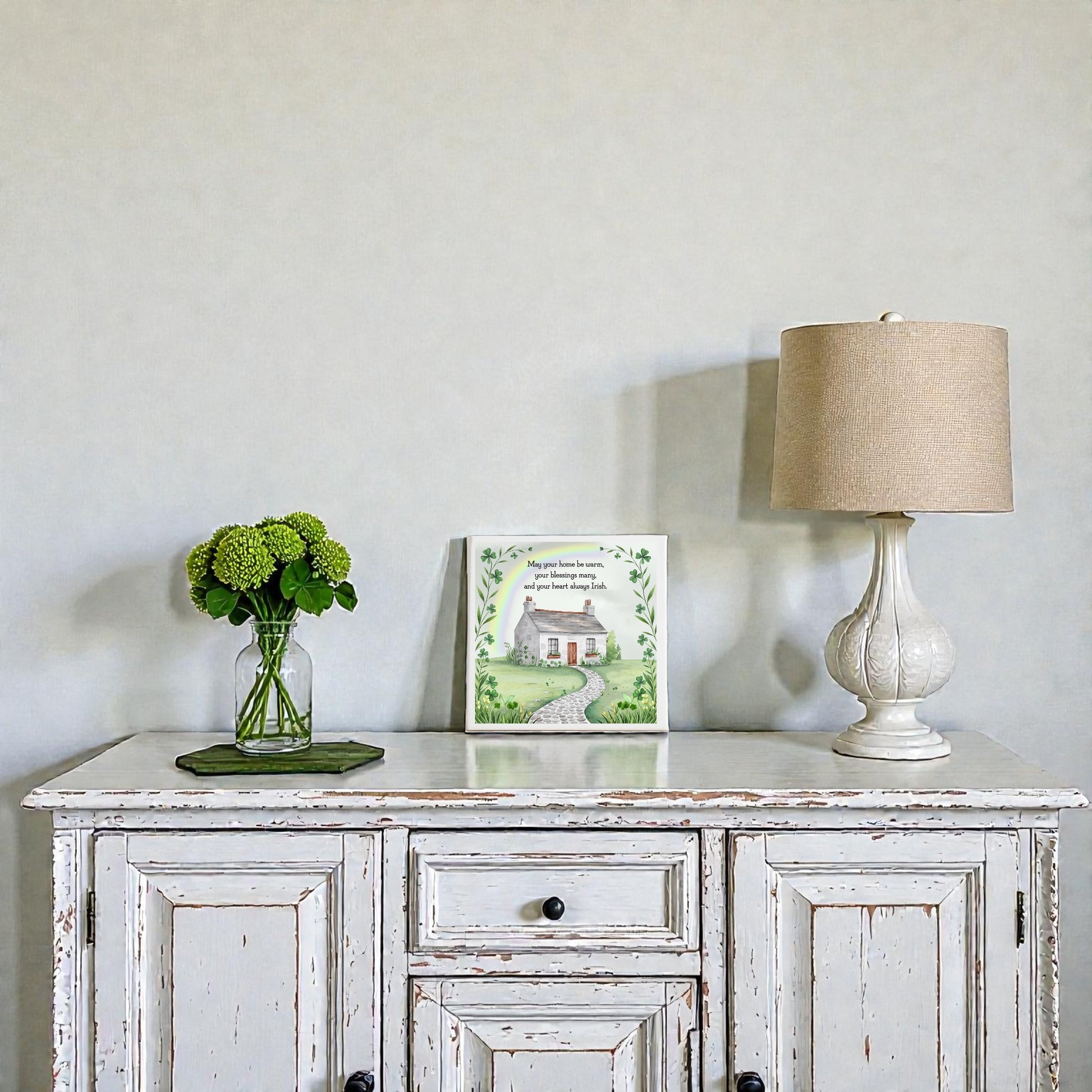 A white distressed wooden sideboard displays the Mirabilia Boutique Irish Heart Home Blessing Canvas – 8x8 Quote Sign, a clear vase with green flowers, and a beige lamp—perfect housewarming decor against a plain light gray wall.