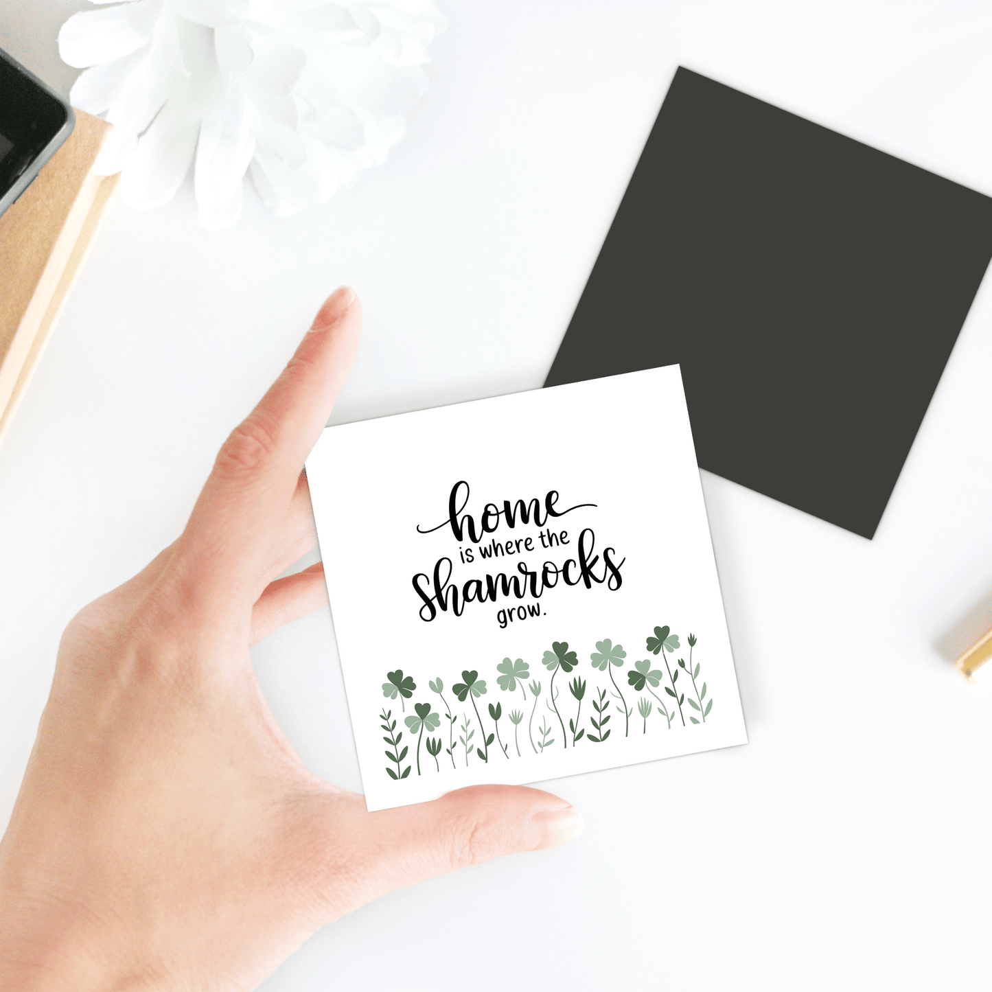 A hand holds the “Home Is Where the Shamrocks Grow” magnet by Mirabilia Boutique, featuring green shamrock illustrations—perfect as Irish home decor or a spring fridge accessory. Scene includes a black square, white flower, and gold pen.