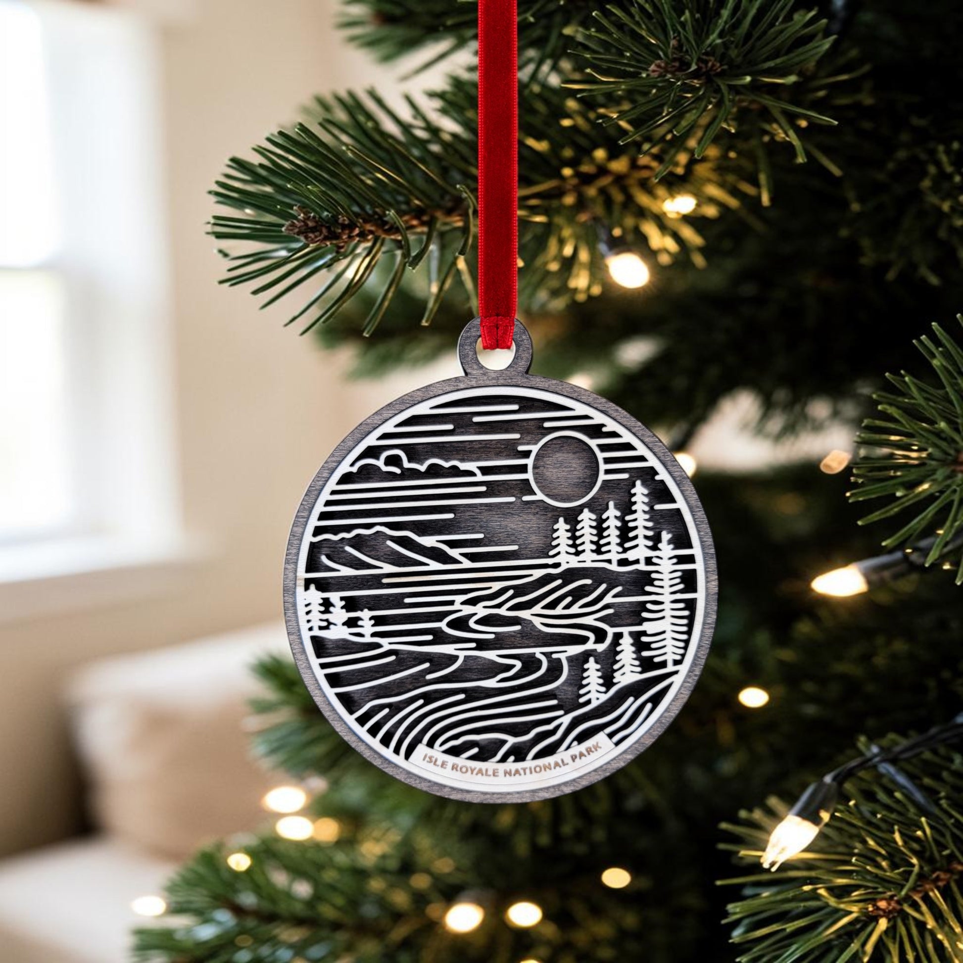 The Isle Royale National Park Line Art Christmas Ornament by Mirabilia Boutique, featuring a scenic line art design of the park nestled in Lake Superior, hangs from a red ribbon on a Christmas tree surrounded by glowing string lights.