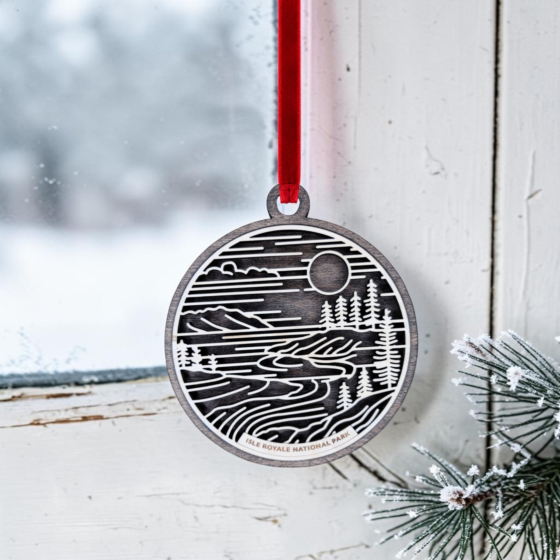 A silver Isle Royale National Park Line Art Christmas Ornament by Mirabilia Boutique hangs by a red ribbon on a window. Pine needles dusted with snow are nearby, and through the glass, a snowy scene overlooking Lake Superior is visible outside. The ornament features a landscape design and reads "Isle Royale National Park.