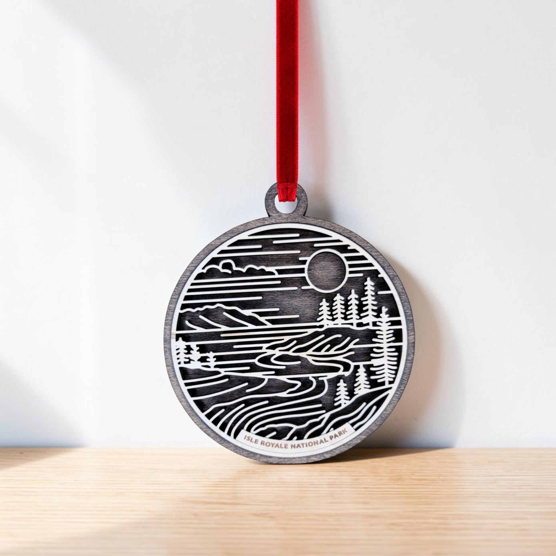 The Isle Royale National Park Line Art Christmas Ornament by Mirabilia Boutique—a round wooden ornament featuring a black and white landscape with trees, water, and the sun inspired by Isle Royale National Park on Lake Superior—hangs from a red ribbon against a white wall, resting on a light wooden surface.