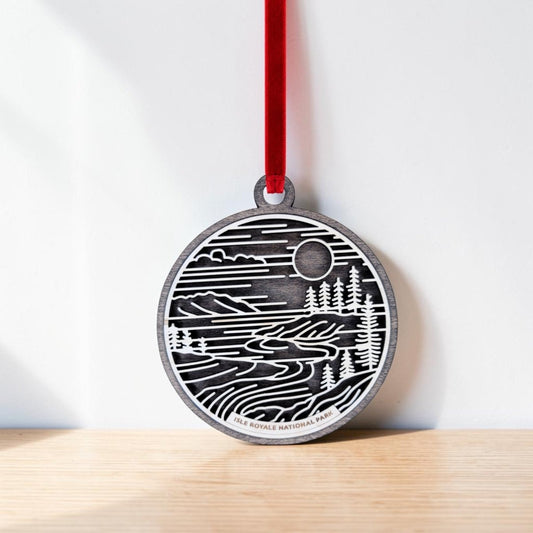 The Isle Royale National Park Line Art Christmas Ornament by Mirabilia Boutique—a round wooden ornament featuring a black and white landscape with trees, water, and the sun inspired by Isle Royale National Park on Lake Superior—hangs from a red ribbon against a white wall, resting on a light wooden surface.