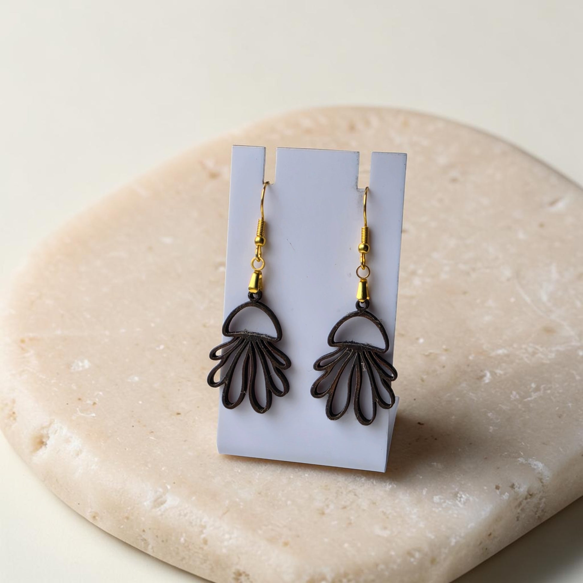 A pair of Jellyfish Earrings by Mirabilia Boutique, crafted from lasercut walnut wood, featuring a fan-shaped design, are showcased on a white card stand atop a beige stone surface.