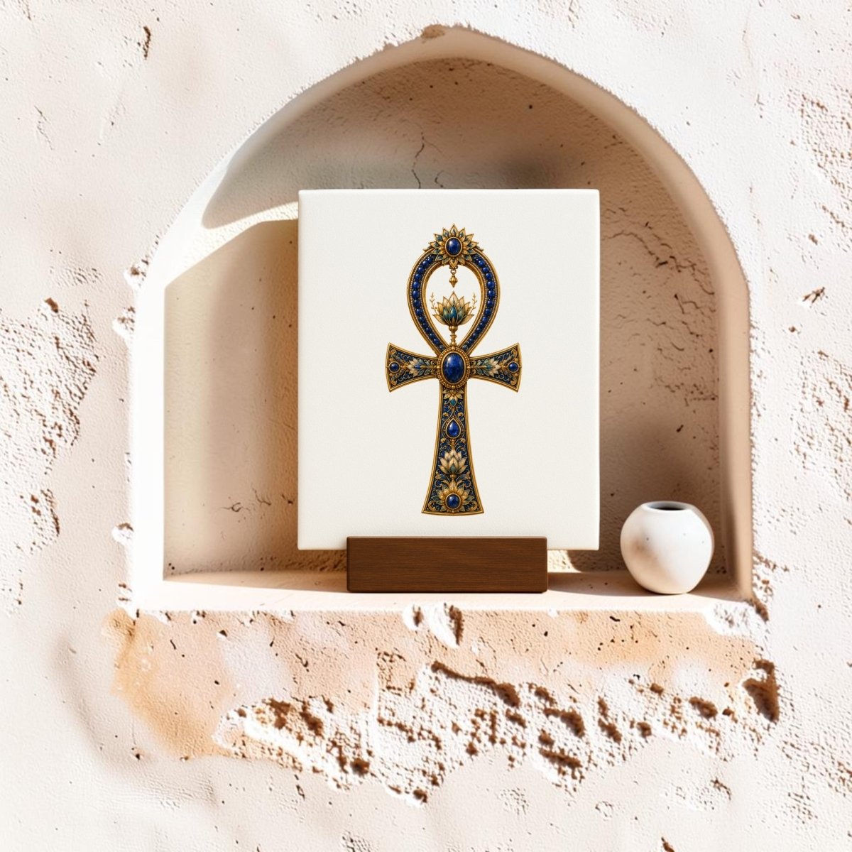 The Jeweled Ankh Mini Sign by Mirabilia Boutique, inspired by Ancient Egyptian decor and adorned with lapis and gold accents, is displayed on a wooden stand within a textured wall niche, accompanied by a small round white vase.