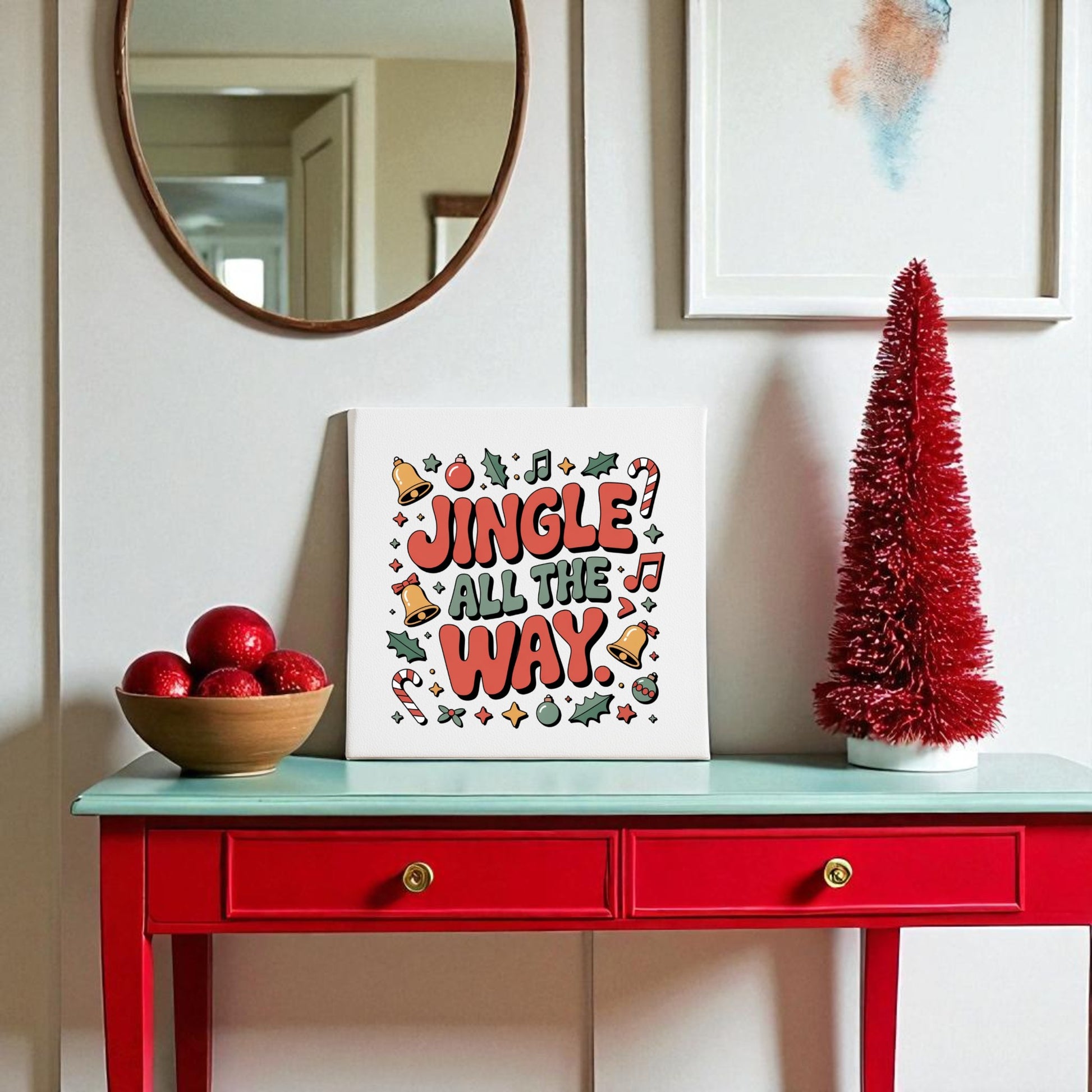 A red console table displays a bowl of red ornaments, a Mirabilia Boutique "Jingle All the Way Canvas" 8x8 wall art, and a red Christmas tree decor, all beneath a round mirror for festive holiday flair.