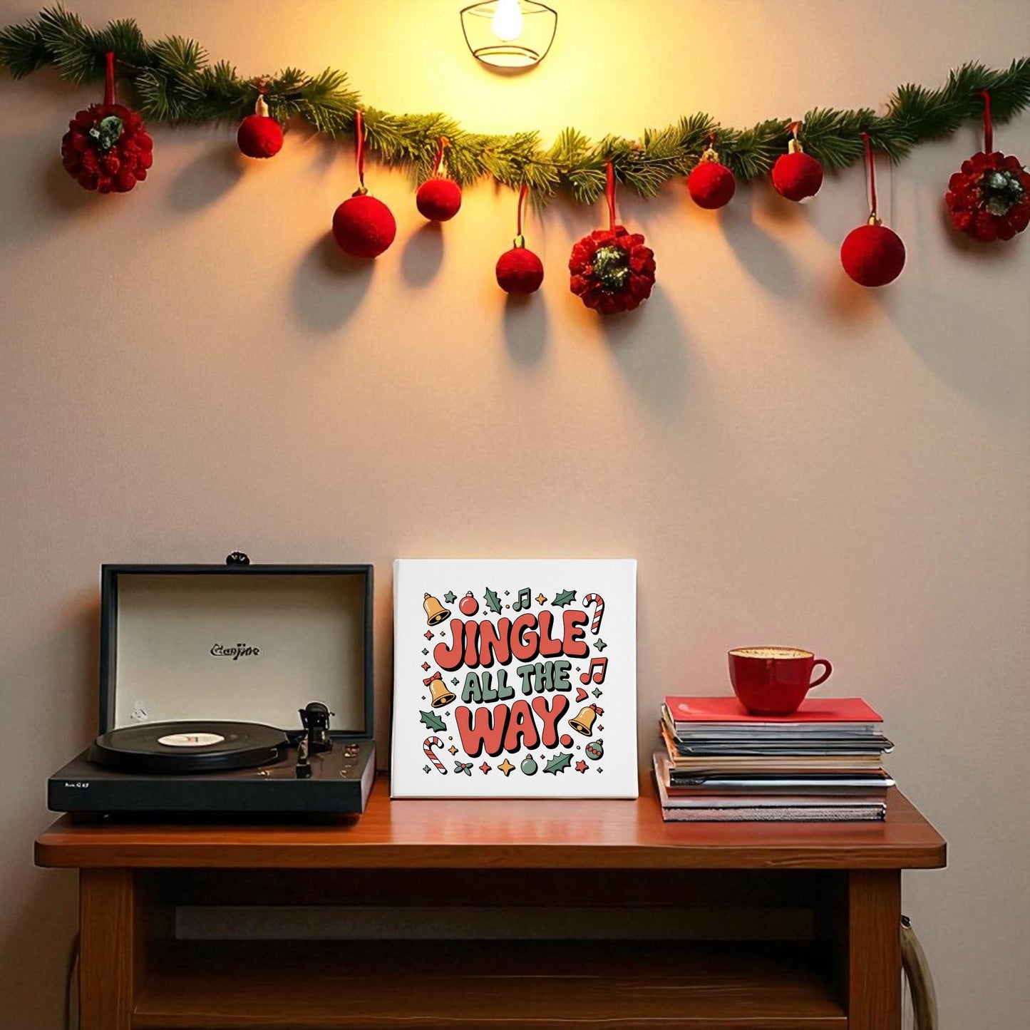 A festive table shows a record player, records with a red cup, and Mirabilia Boutique’s "Jingle All the Way Canvas" 8x8 wall art. Above, pine garland and red ornaments decorate the wall, softly lit by a warm sconce.