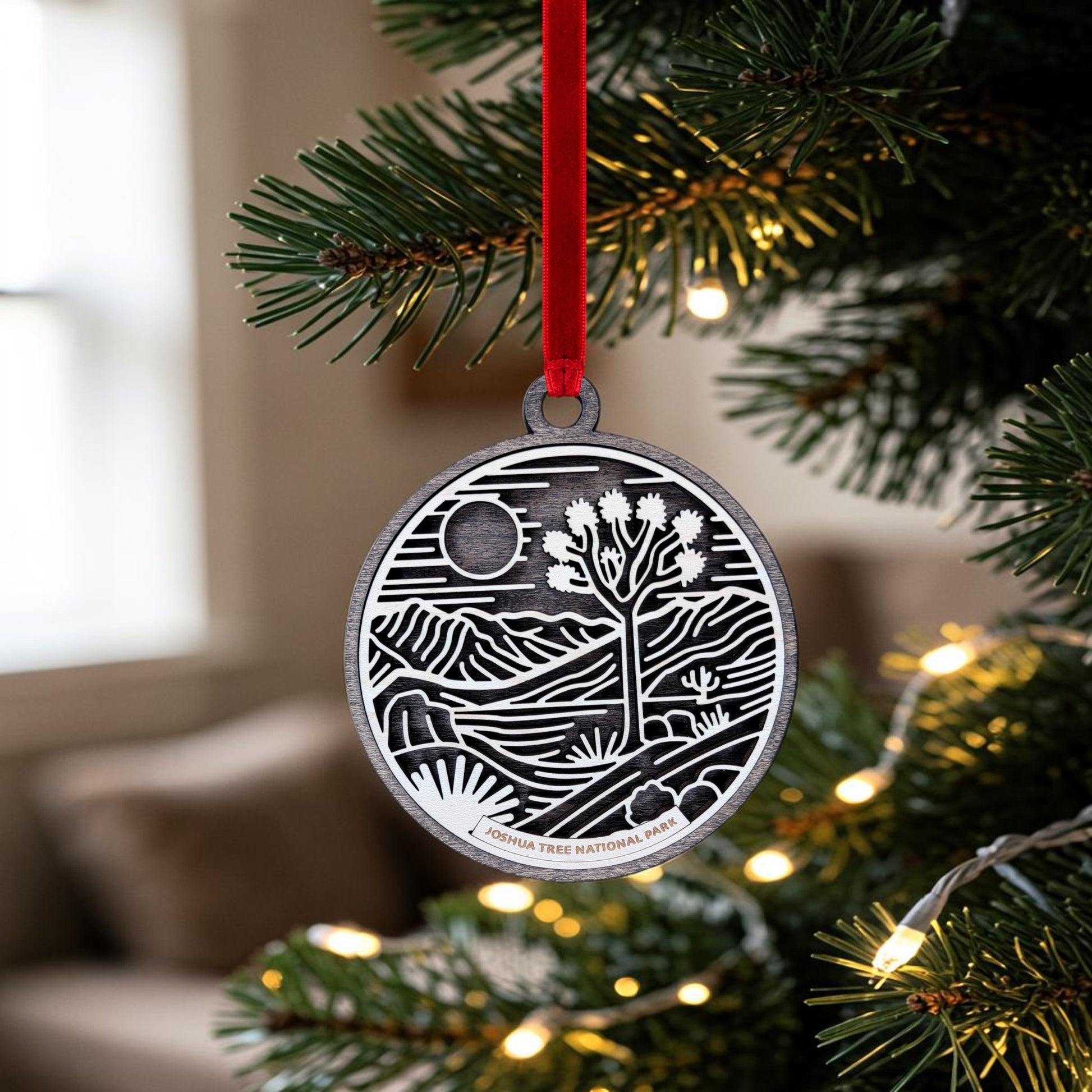 The Joshua Tree National Park Line Art Christmas Ornament by Mirabilia Boutique, featuring an oval wooden design with a desert landscape and a Joshua tree, hangs from a red ribbon amid green branches and white string lights on the Christmas tree. Inspired by the beauty of Joshua Tree National Park, this unique ornament brings a touch of Southwest charm to your holiday decor.