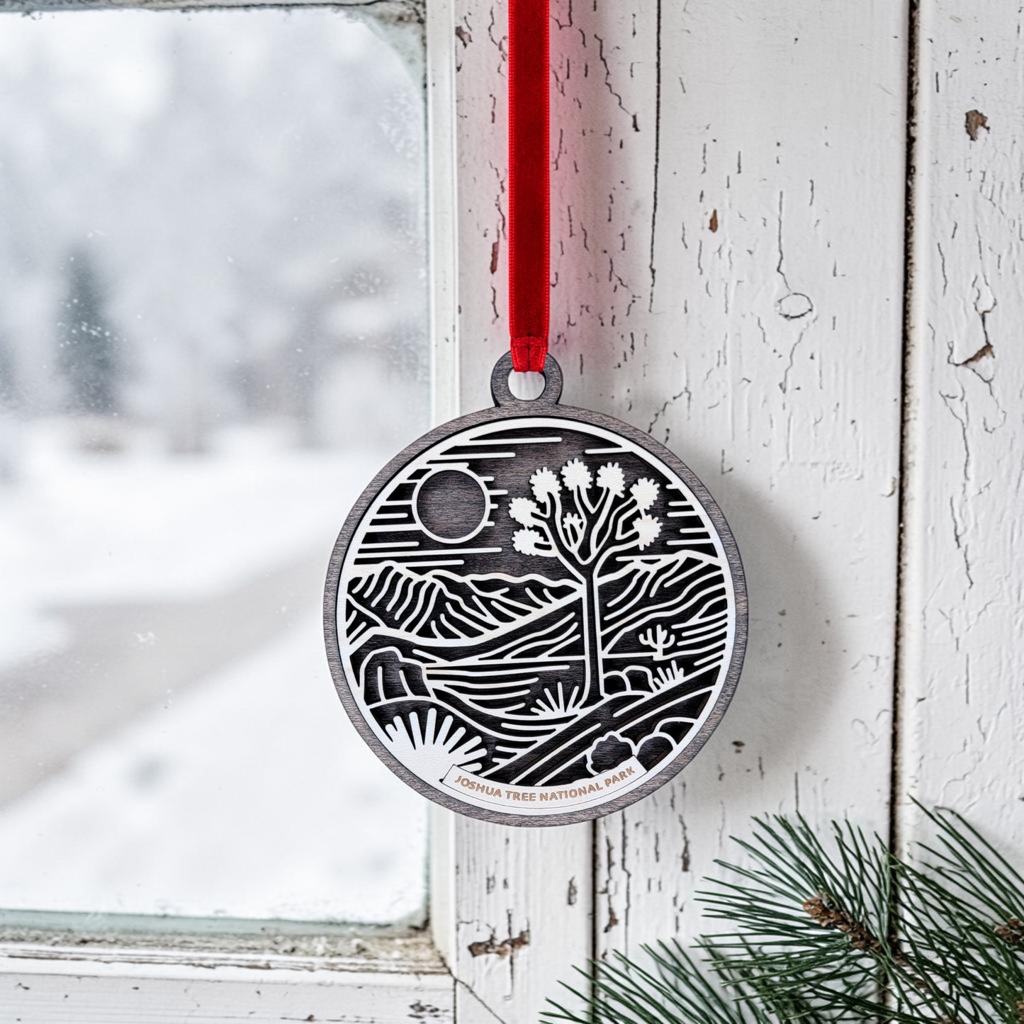 The Joshua Tree National Park Line Art Christmas Ornament by Mirabilia Boutique—a round wooden ornament featuring a desert landscape—hangs by a red ribbon on a white, weathered window frame next to green pine needles, with snowy scenery visible outside.
