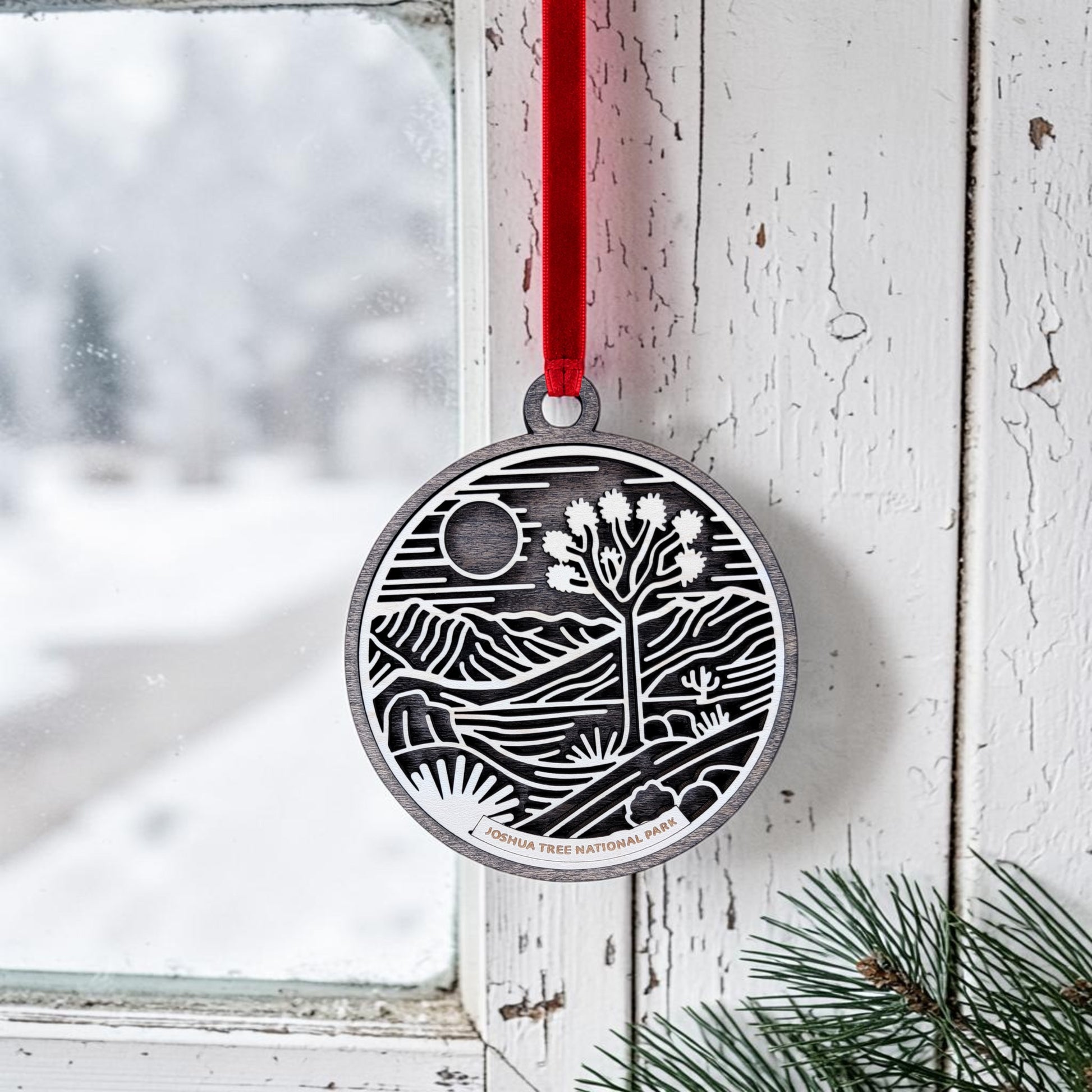 The Joshua Tree National Park Line Art Christmas Ornament by Mirabilia Boutique—a round wooden ornament featuring a desert landscape—hangs by a red ribbon on a white, weathered window frame next to green pine needles, with snowy scenery visible outside.