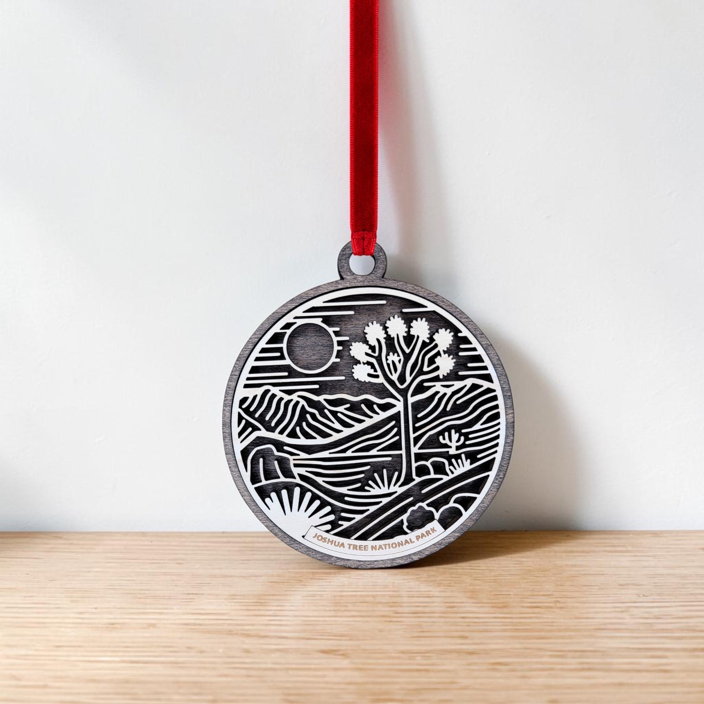 The Joshua Tree National Park Line Art Christmas Ornament by Mirabilia Boutique hangs from a red ribbon against a white wall and wooden surface, showcasing a round wooden design featuring the iconic Joshua tree, sun, and mountains in a desert landscape.