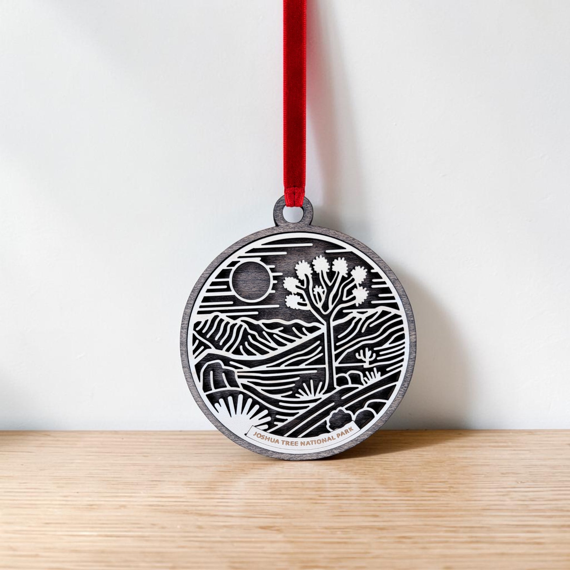 The Joshua Tree National Park Line Art Christmas Ornament by Mirabilia Boutique hangs from a red ribbon against a white wall and wooden surface, showcasing a round wooden design featuring the iconic Joshua tree, sun, and mountains in a desert landscape.