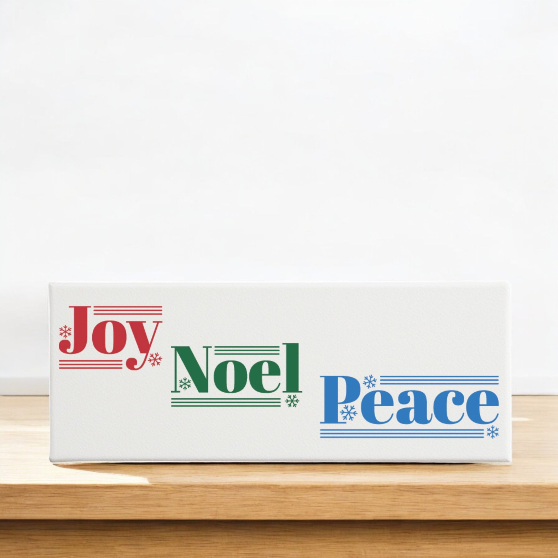 The Mirabilia Boutique Joy Noel Peace Christmas Sign is a white 4x12 canvas shelf sitter featuring "Joy" in red, "Noel" in green, and "Peace" in blue with festive lines and snowflakes—perfect holiday decor for any space.