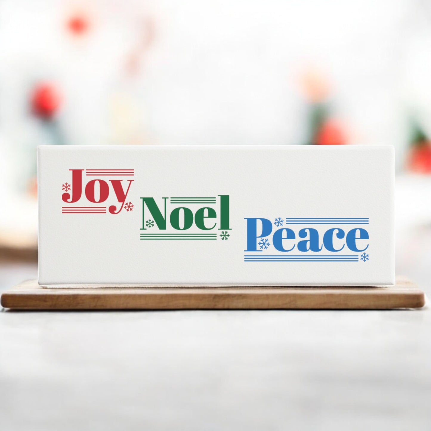 The Joy Noel Peace Christmas Sign by Mirabilia Boutique is a 4x12 canvas shelf sitter featuring festive red, green, and blue text with snowflakes and musical staff accents—a charming holiday décor piece for your home.