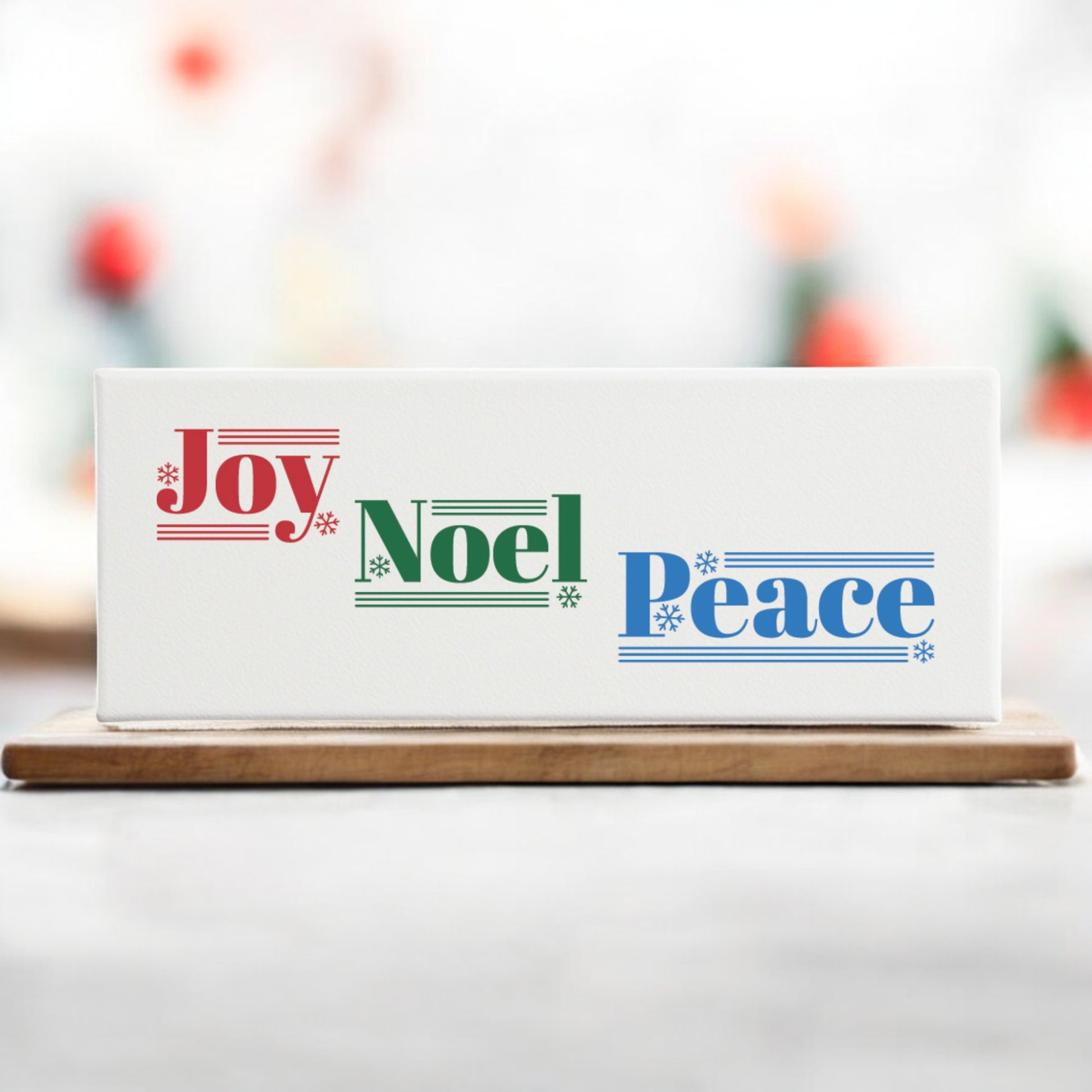 The Joy Noel Peace Christmas Sign by Mirabilia Boutique is a 4x12 canvas shelf sitter featuring festive red, green, and blue text with snowflakes and musical staff accents—a charming holiday décor piece for your home.