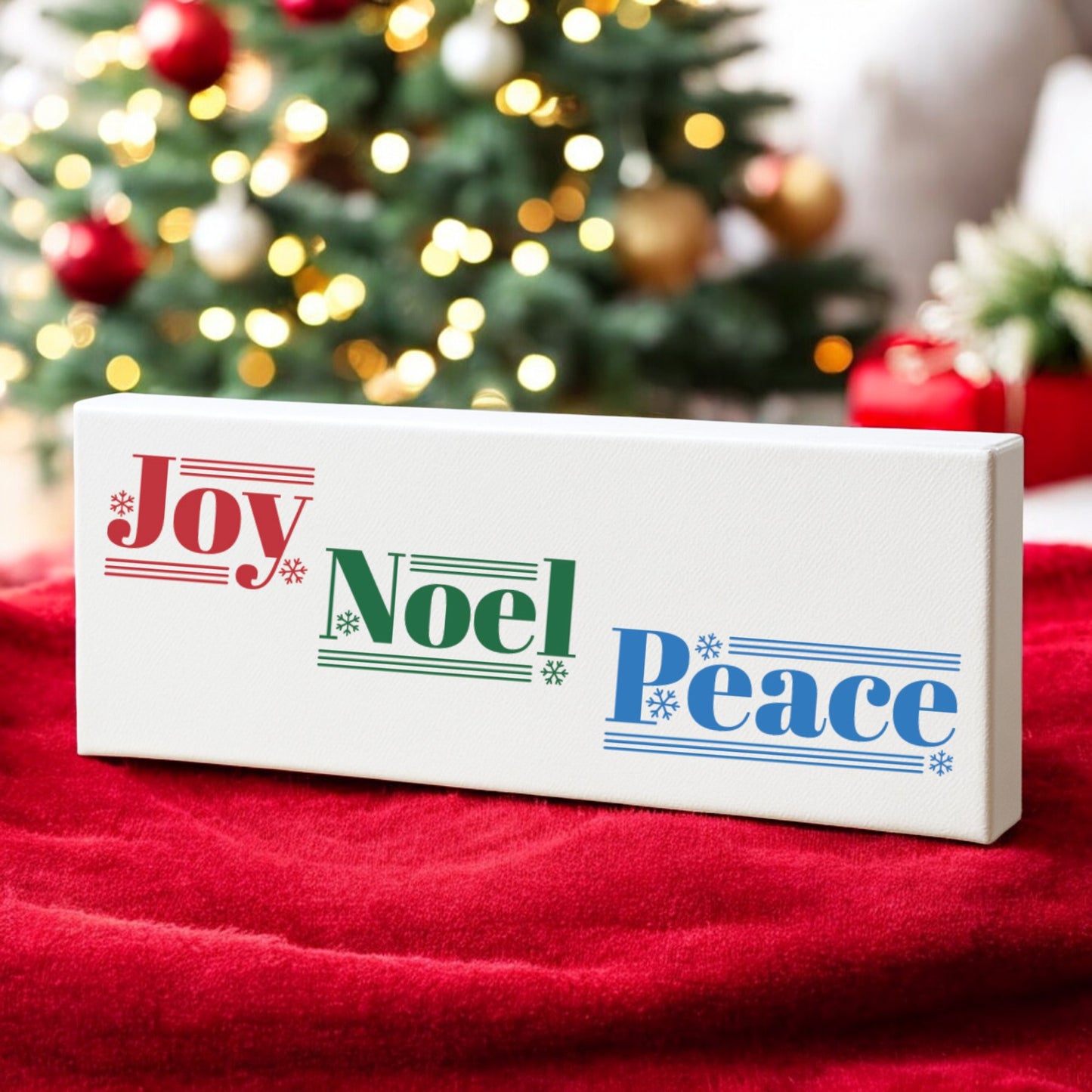 The Mirabilia Boutique Joy Noel Peace Christmas Sign, a 4x12 white canvas shelf sitter with festive lettering, rests on a red blanket amid holiday décor and a softly blurred Christmas tree in the background.