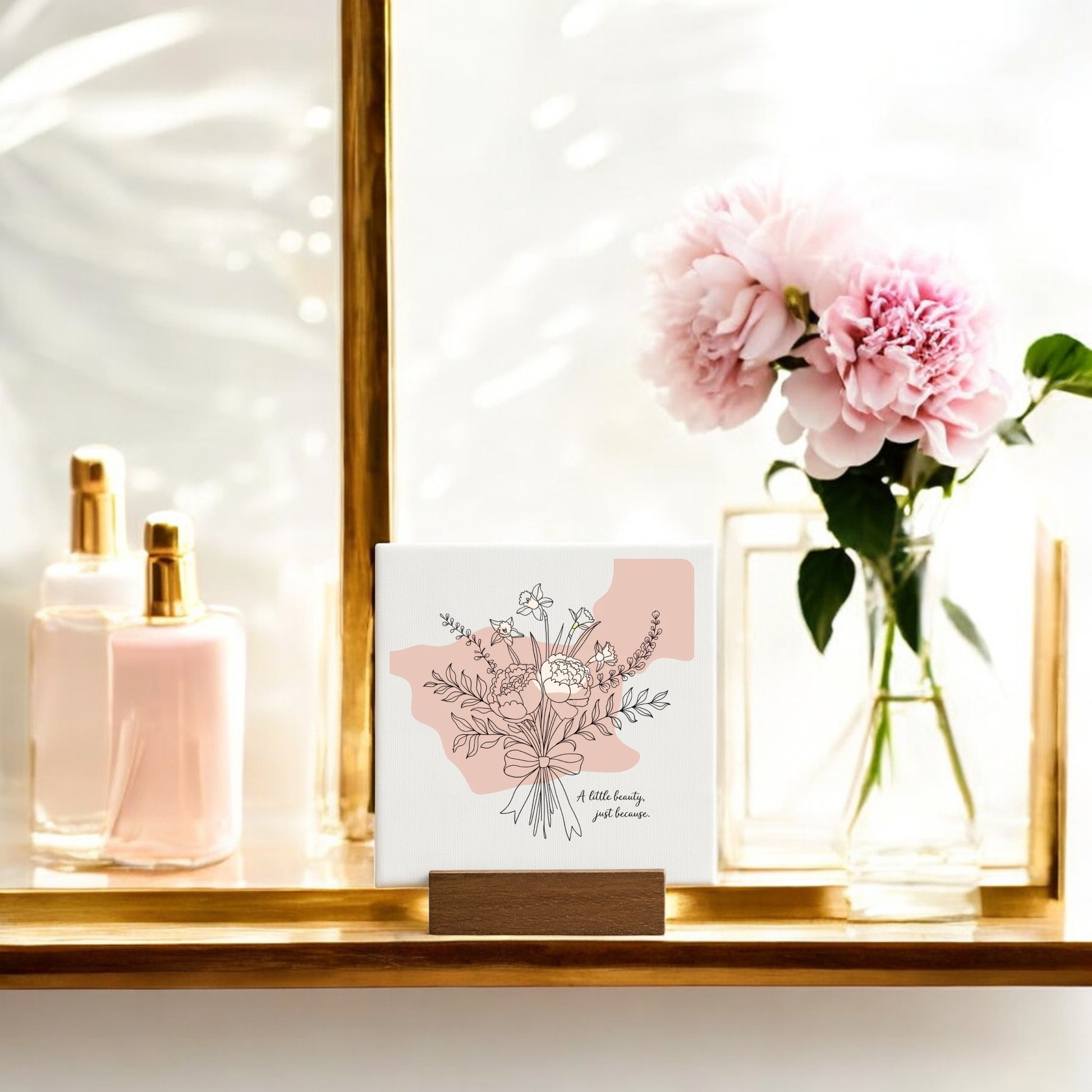 A softly lit shelf displays Mirabilia Boutique’s “A Little Beauty Mini Canvas – Just Because Floral Sign,” accented with pink peonies and perfume bottles—a charming, minimalist decor piece or thoughtful gift.