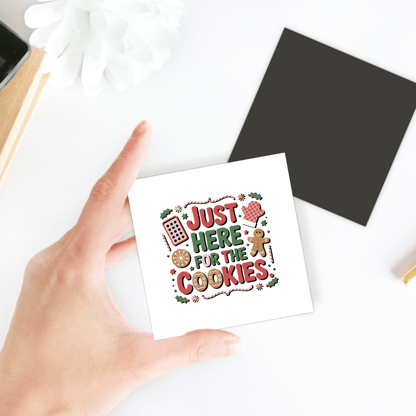 A hand holds the "Just Here for the Cookies" Magnet by Mirabilia Boutique, featuring cookie and gingerbread designs—a funny Christmas fridge decor perfect for cookie lovers, with a black square and white flower in the holiday kitchen background.