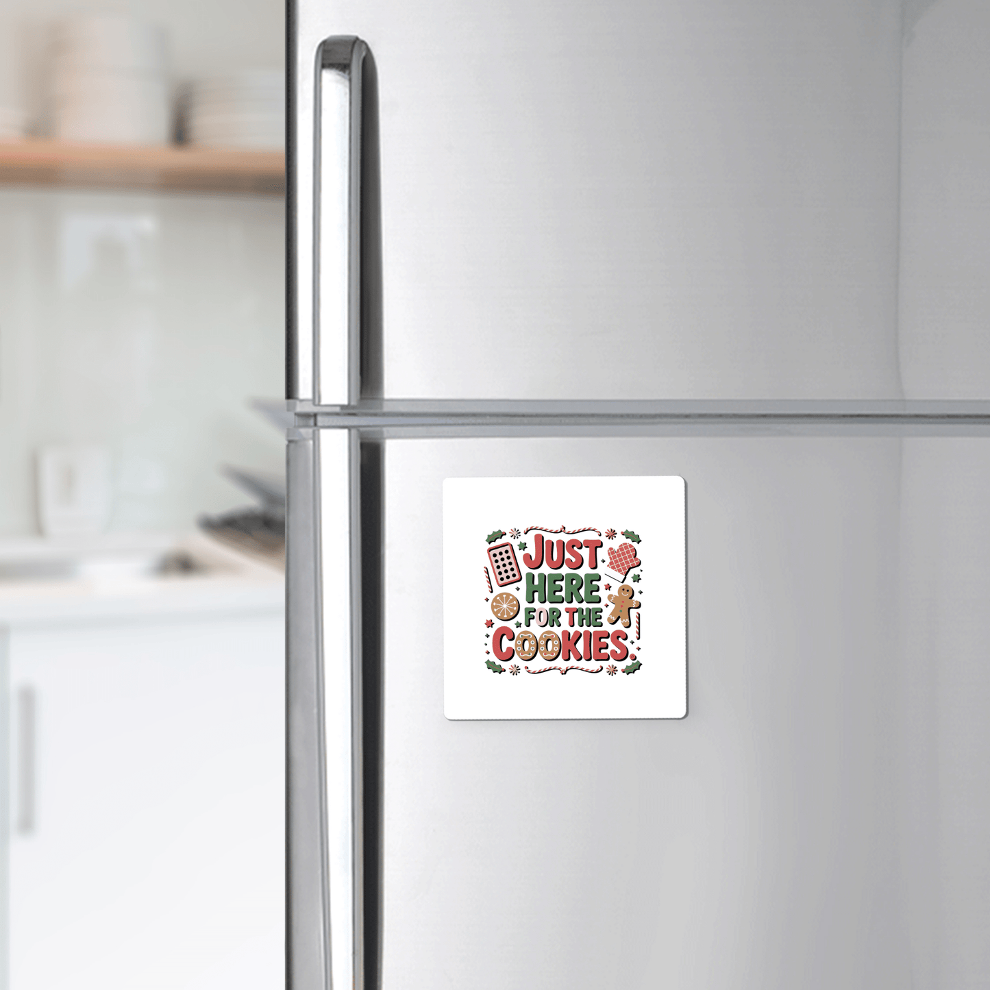A stainless steel fridge is decorated with the Mirabilia Boutique "Just Here for the Cookies Magnet"—a funny Christmas fridge decor that's perfect for holiday kitchens and cookie lovers. Kitchen counter and shelves are visible in the background.