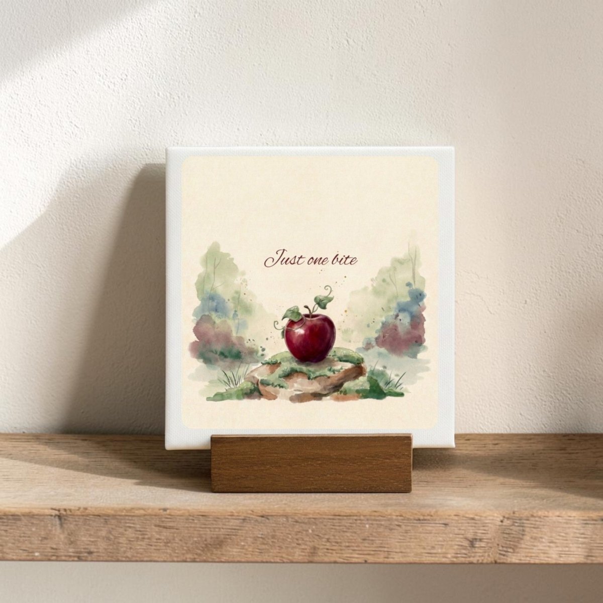 The Just One Bite Mini Canvas Sign by Mirabilia Boutique features a red apple with green leaves and the phrase "Just one bite" above it, surrounded by watercolor-style foliage on a wooden shelf—making it a charming fairy tale decor accent for any woodland-inspired space.