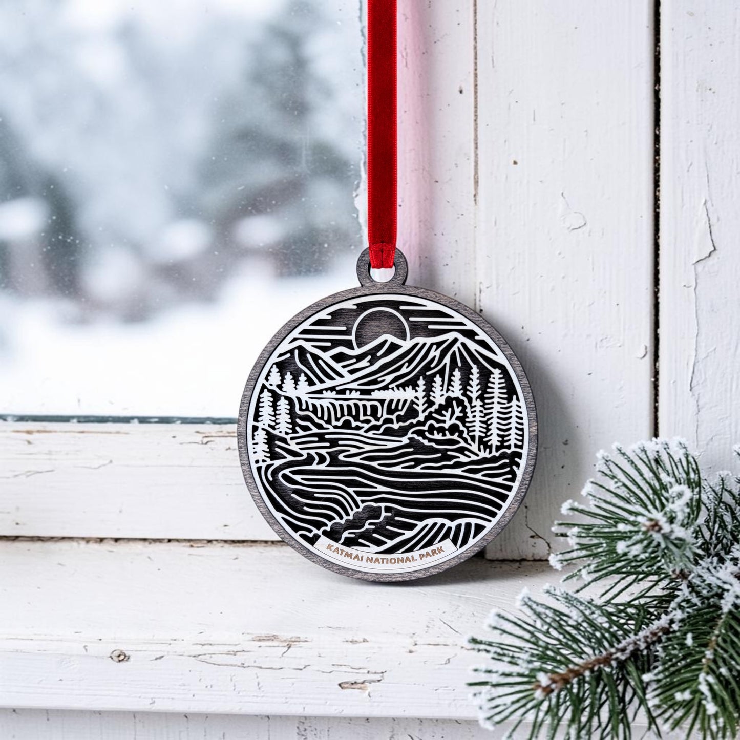 A Katmai National Park Line Art Christmas Ornament by Mirabilia Boutique, showcasing a handcrafted wooden design with a nature-inspired scene and "Katmai National Park" text, hangs from a red ribbon on a snowy window frame. Frosted pine branches in the corner evoke the wild beauty of Alaska brown bears and their pristine habitat.