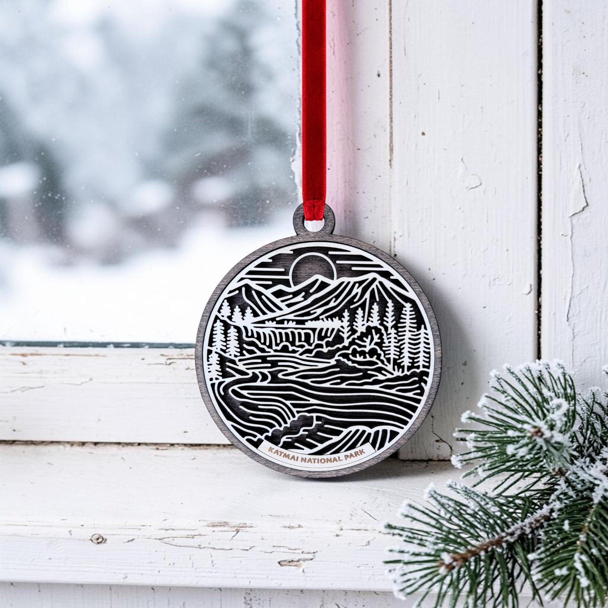 A Katmai National Park Line Art Christmas Ornament by Mirabilia Boutique, showcasing a handcrafted wooden design with a nature-inspired scene and "Katmai National Park" text, hangs from a red ribbon on a snowy window frame. Frosted pine branches in the corner evoke the wild beauty of Alaska brown bears and their pristine habitat.