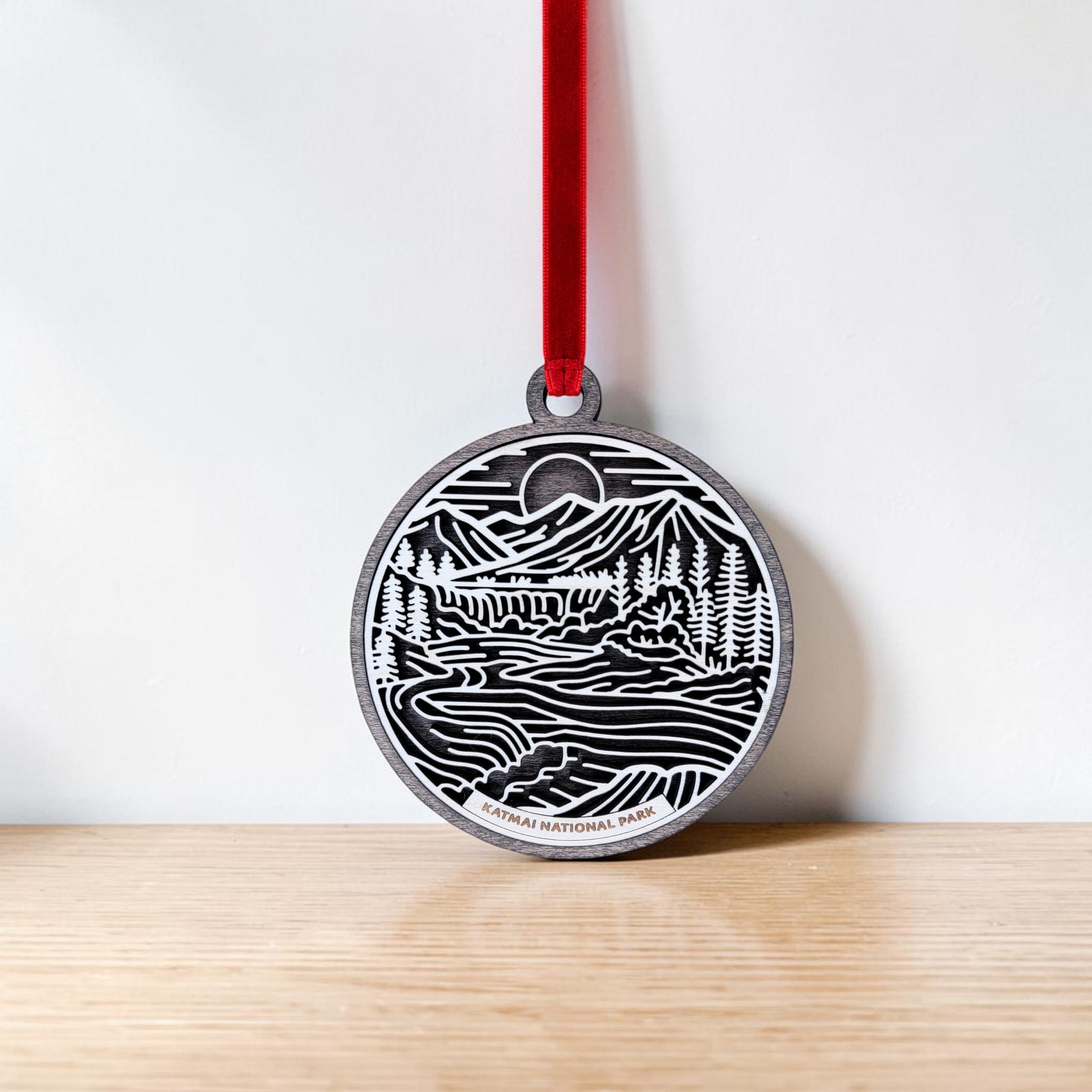 Handcrafted wooden ornament on a red ribbon featuring an engraved line art landscape of mountains, trees, and a river. Text at the bottom reads "KATMAI NATIONAL PARK." The Mirabilia Boutique Katmai National Park Line Art Christmas Ornament is displayed hanging against a white wall above a wooden surface.