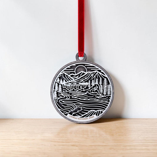Handcrafted wooden ornament on a red ribbon featuring an engraved line art landscape of mountains, trees, and a river. Text at the bottom reads "KATMAI NATIONAL PARK." The Mirabilia Boutique Katmai National Park Line Art Christmas Ornament is displayed hanging against a white wall above a wooden surface.
