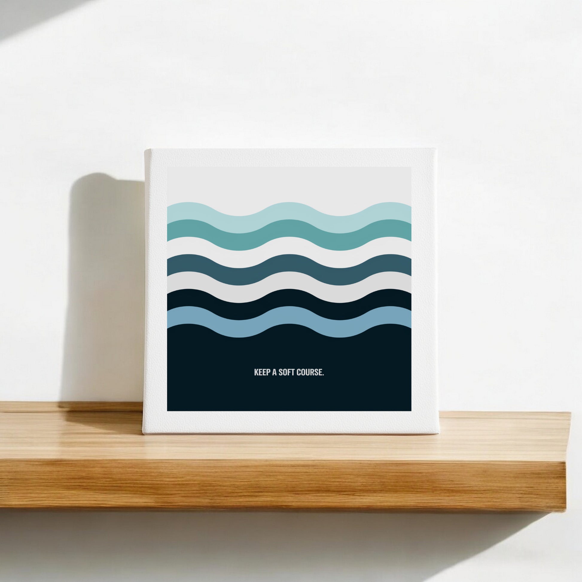 The Mirabilia Boutique "Keep a Soft Course" Canvas Sign (8x8) features minimalist blue and teal ocean waves with calming text, perfect for display on a wooden shelf against a white wall.