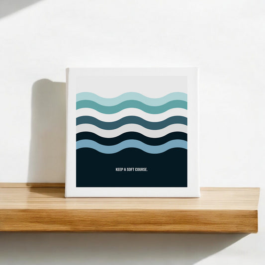 The Mirabilia Boutique "Keep a Soft Course" Canvas Sign (8x8) features minimalist blue and teal ocean waves with calming text, perfect for display on a wooden shelf against a white wall.