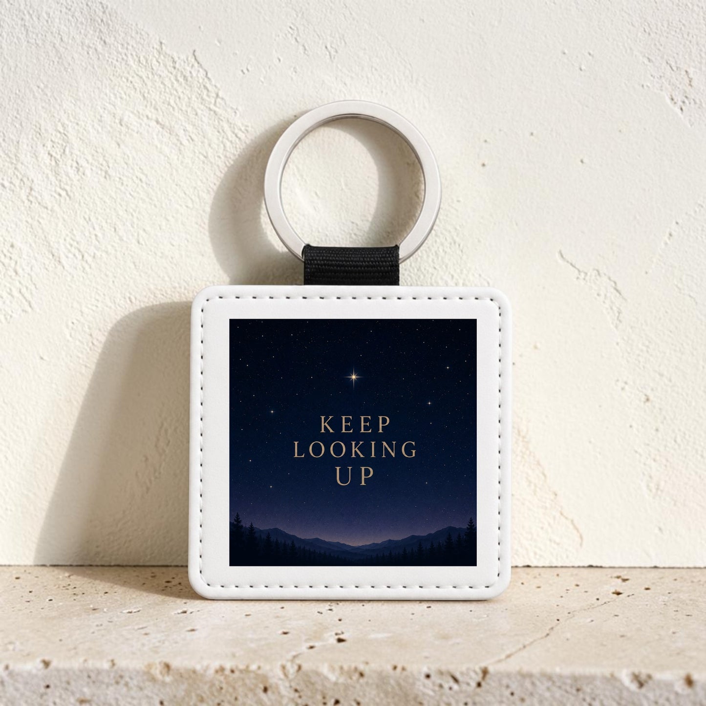 The Keep Looking Up Celestial Keychain by Mirabilia Boutique features a square white design with a night sky and gold “KEEP LOOKING UP” lettering, displayed on a beige textured surface—an inspirational gift to encourage positivity.